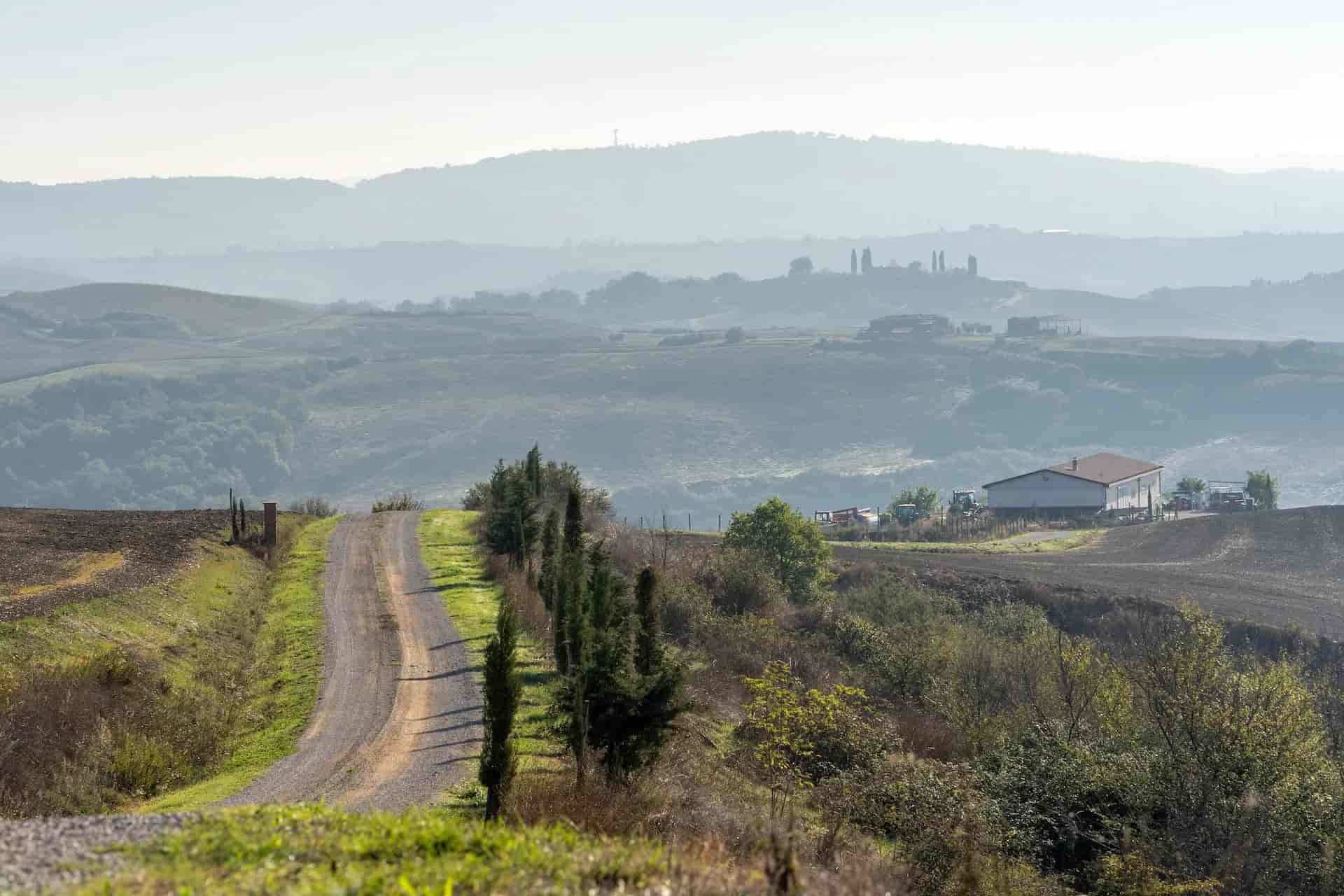 Gravel road winding through rolling hills toward a farm building in hazy Tuscany landscape.