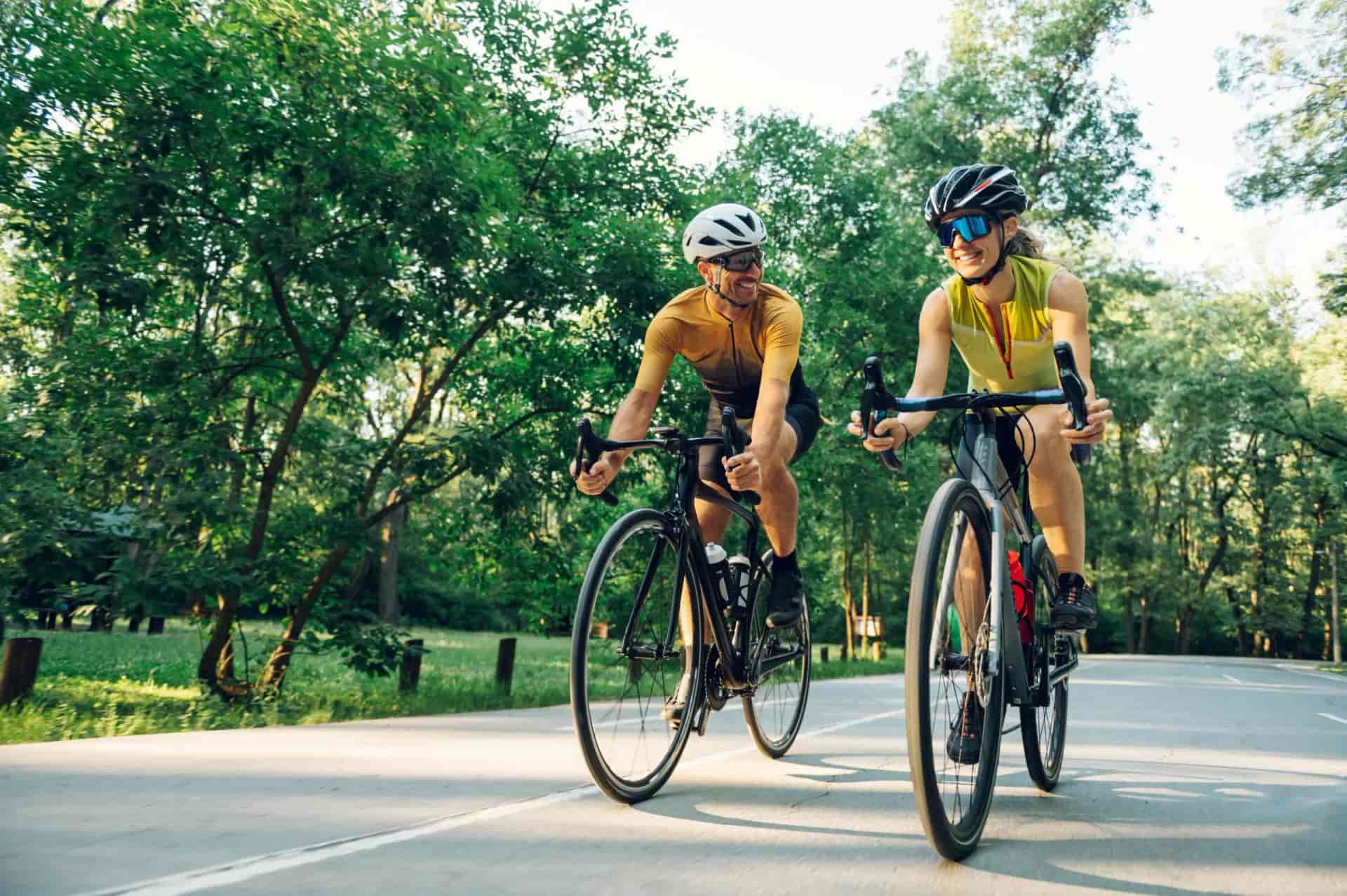 Two cyclists in helmets riding road bikes on a paved path surrounded by green trees.