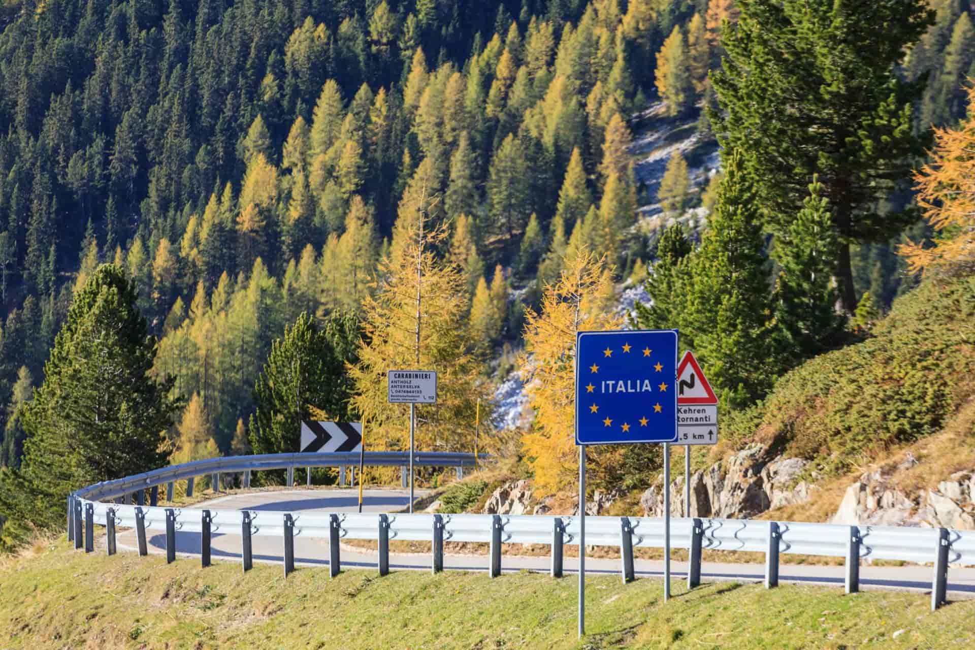 Road sign indicating entry to Italia next to guardrail on mountain road with autumn trees.