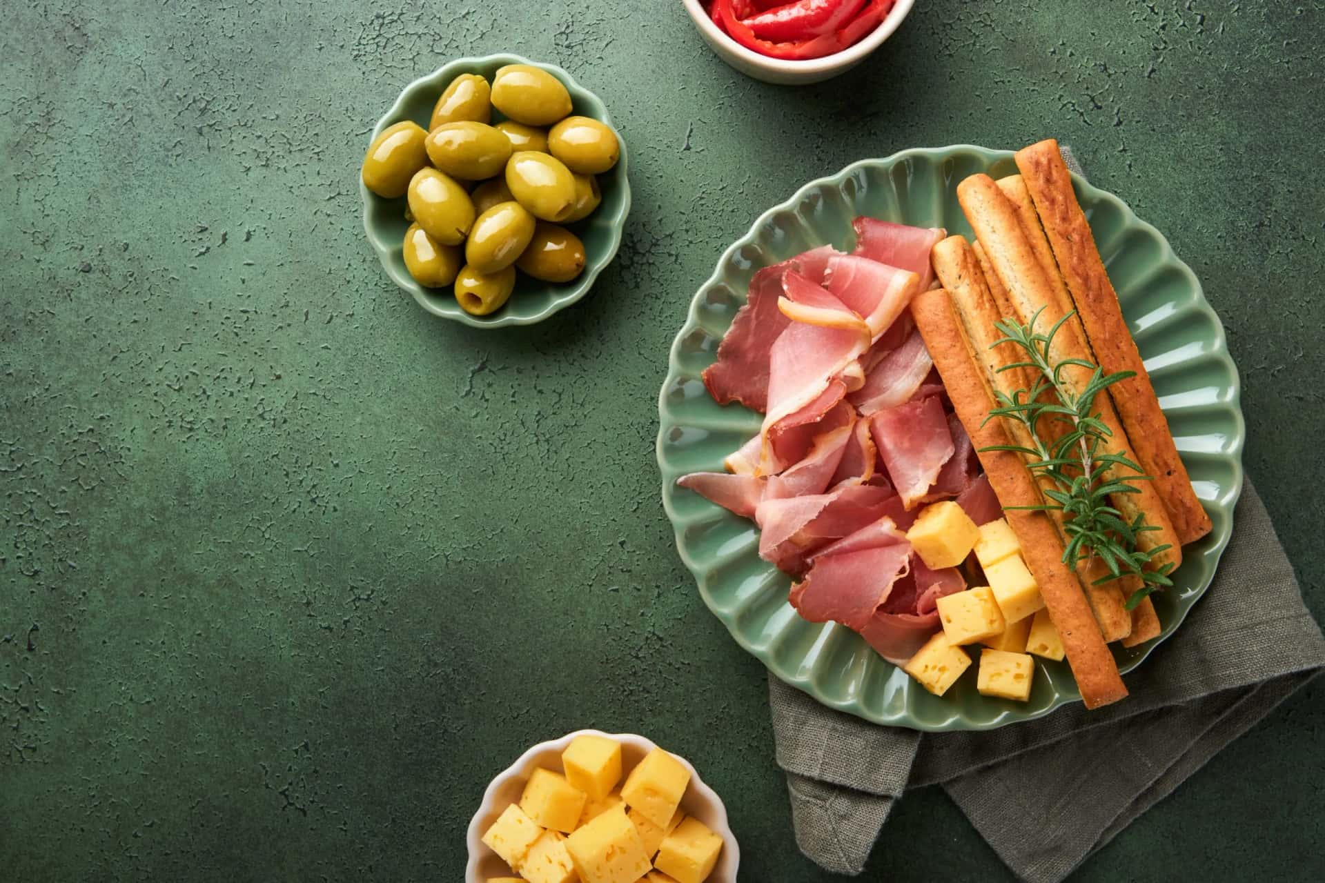 Charcuterie board with prosciutto, cheese cubes, breadsticks, and green olives on a textured green surface.
