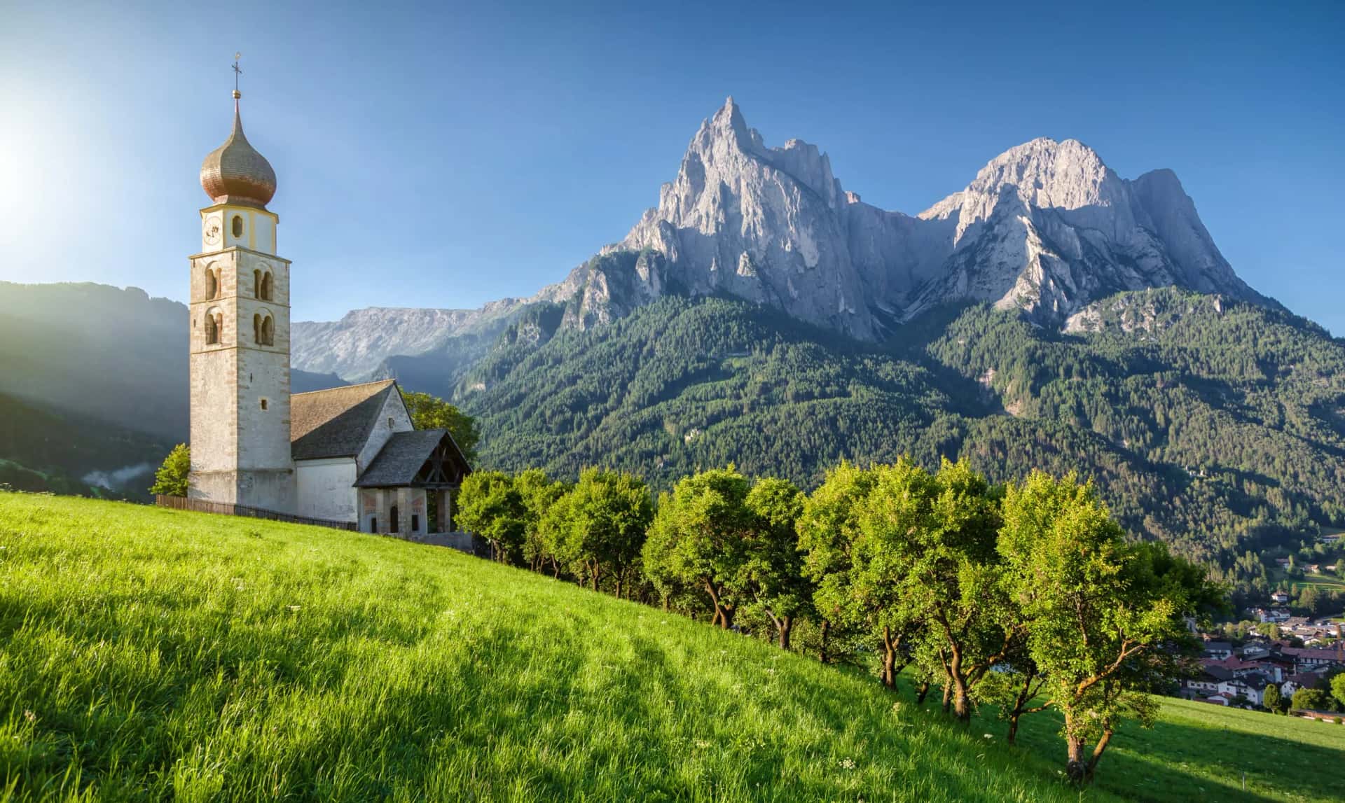 Church with onion dome tower on grassy hill below jagged mountains in the Dolomites