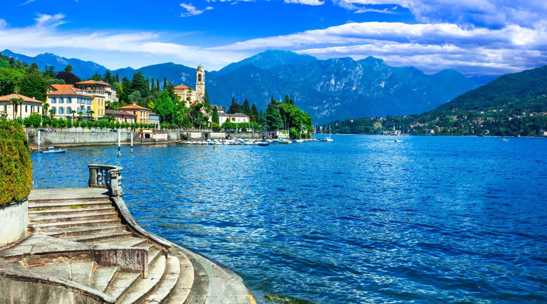 Stone steps leading to deep blue lake water with boats and colorful lakeside town backed by mountains.