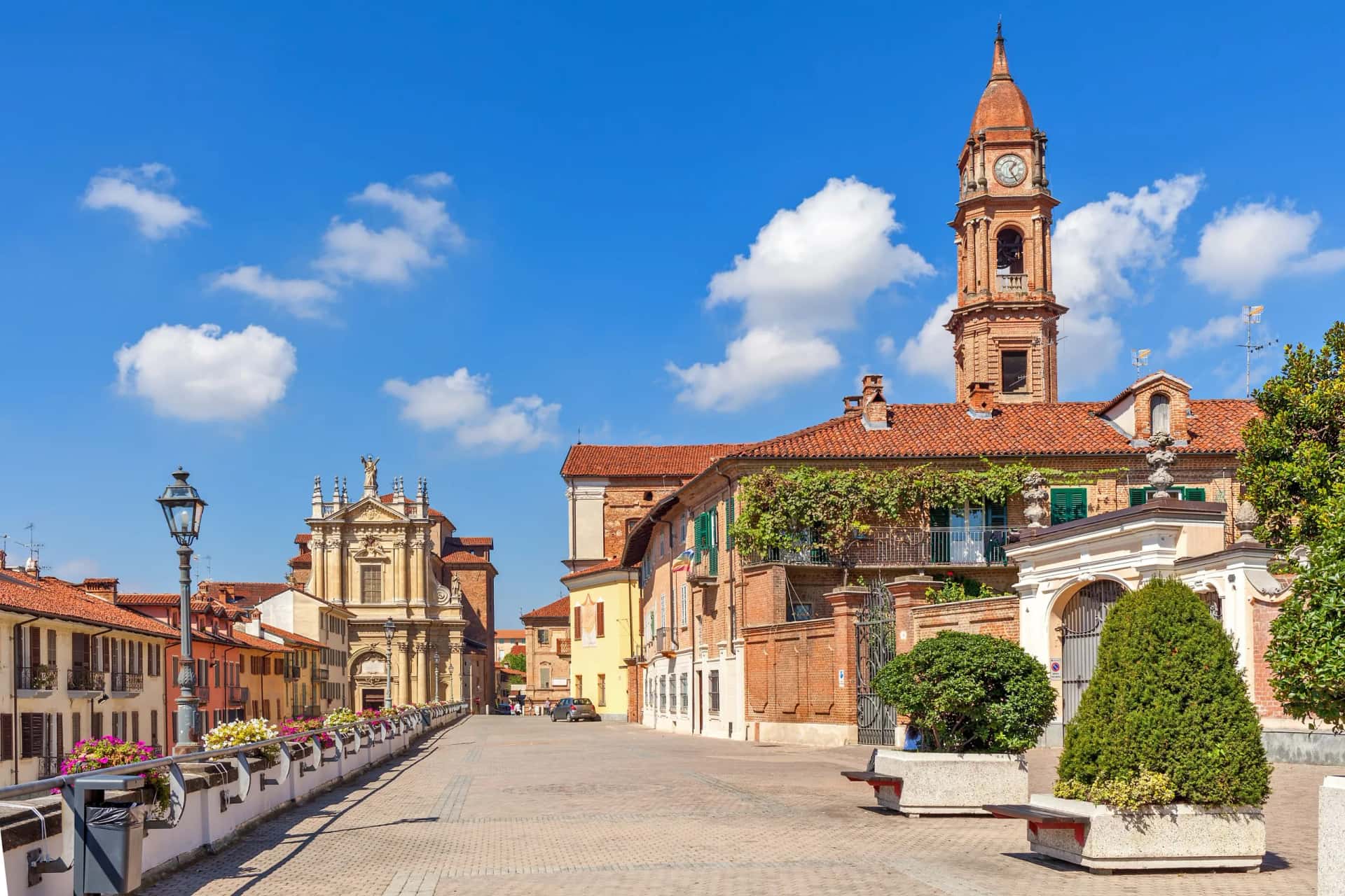 Piedmont town square with Baroque church, brick clock tower, and flower boxes under blue sky.
