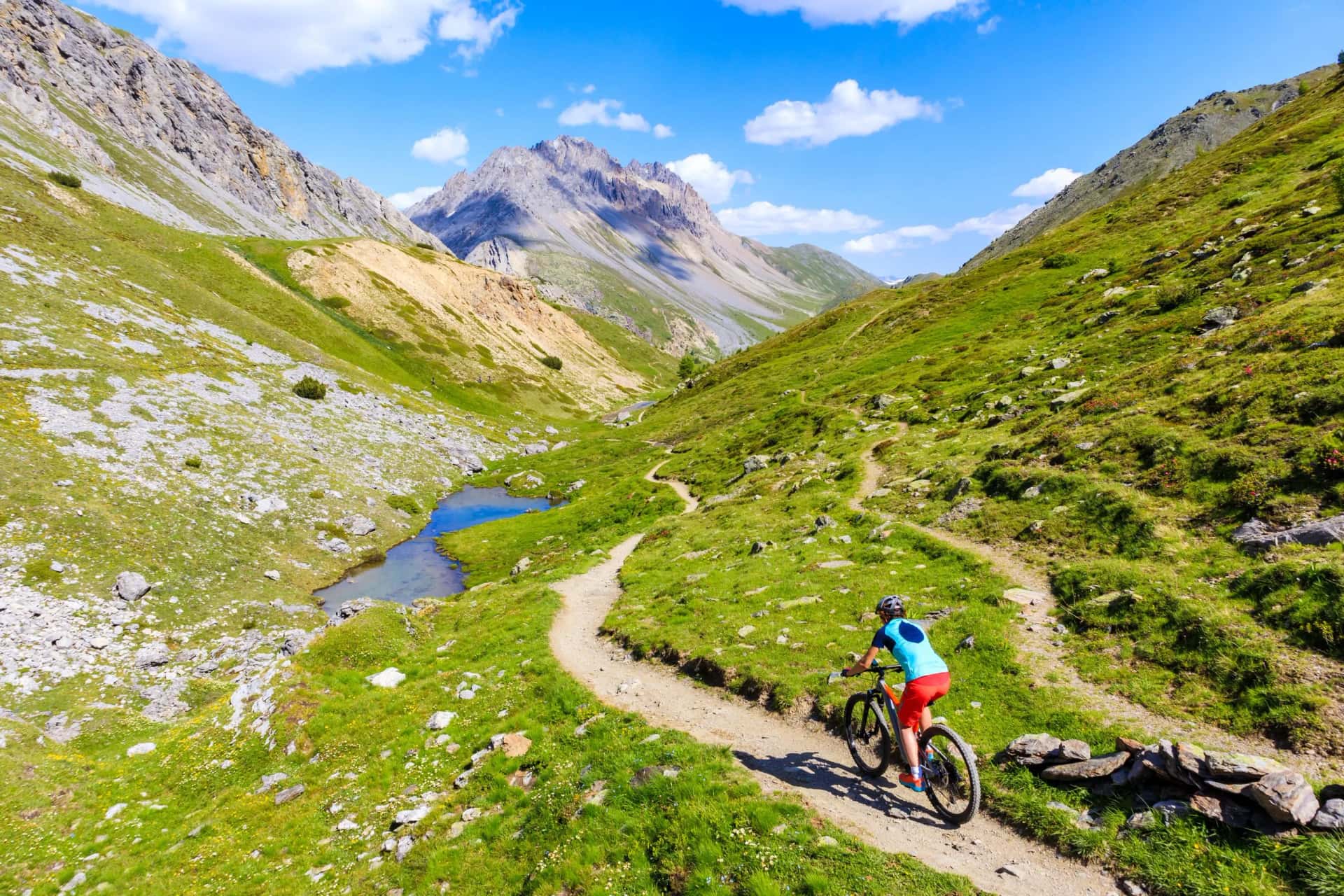 Mountain biking on a dirt trail through green alpine valley with a small stream and rocky peaks.