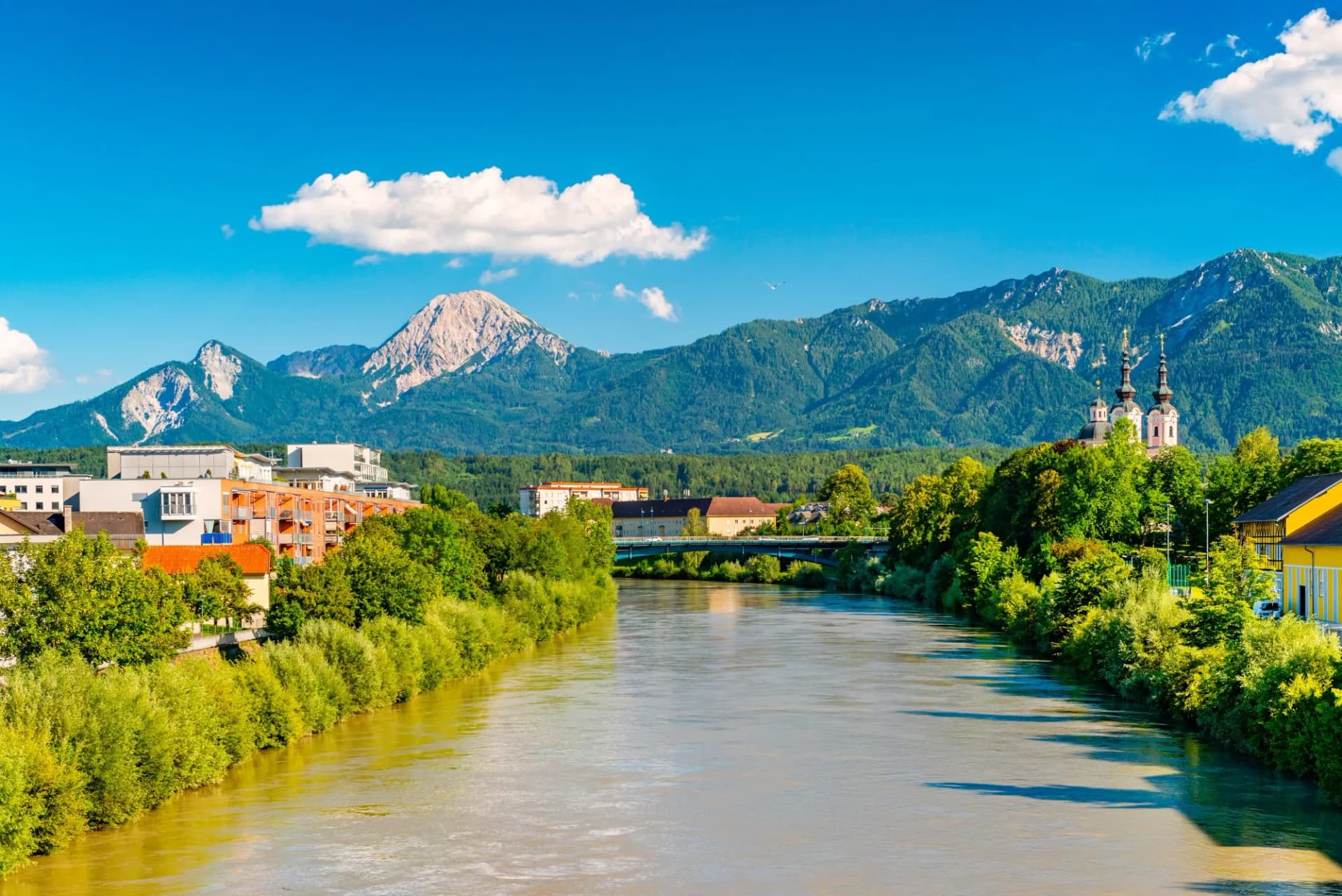 Drau River in Villach with lush green banks, city buildings, and steep mountains under a blue sky.