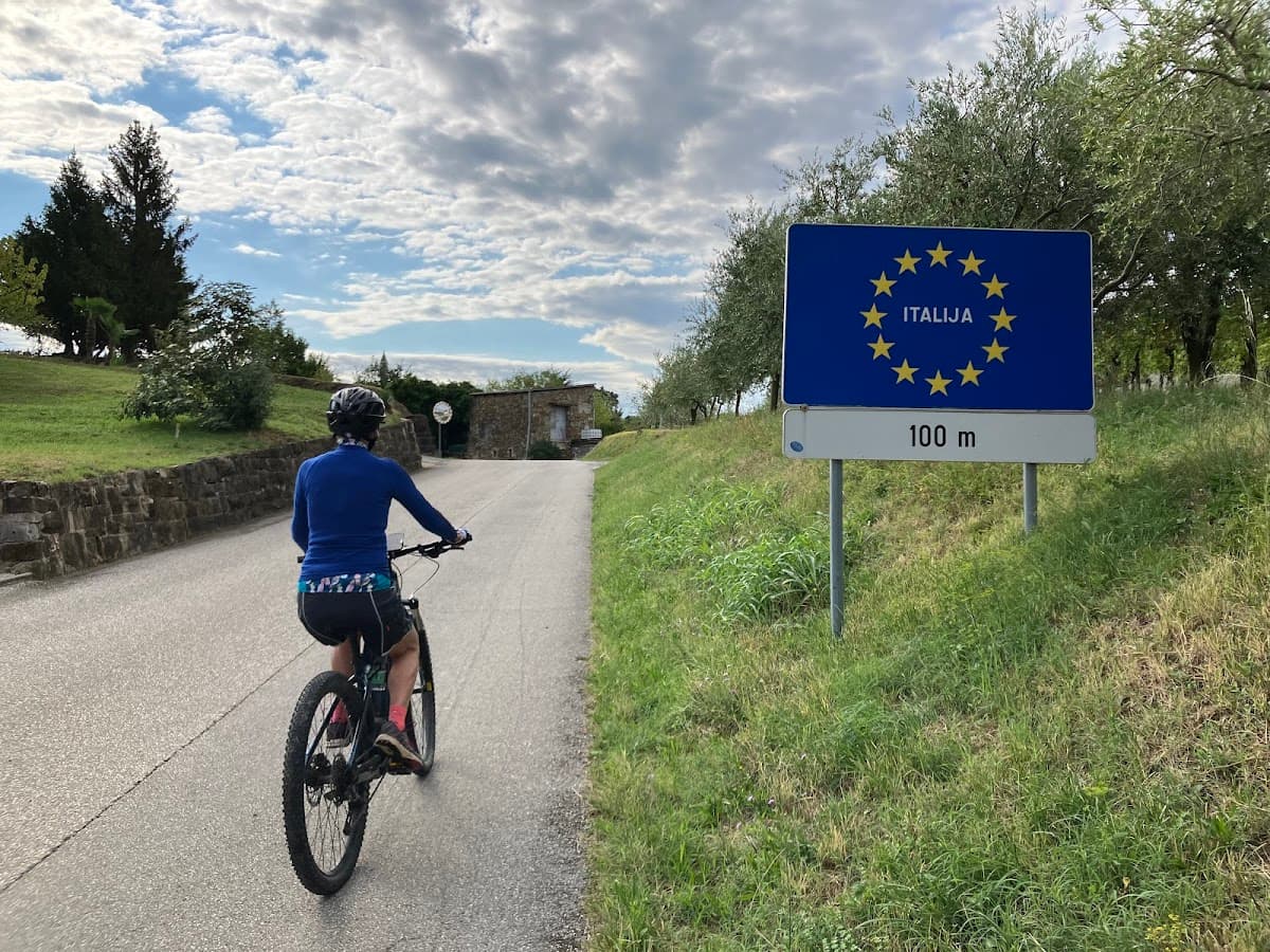Cyclist approaching EU border sign for Italija (Italy) on a paved country road.