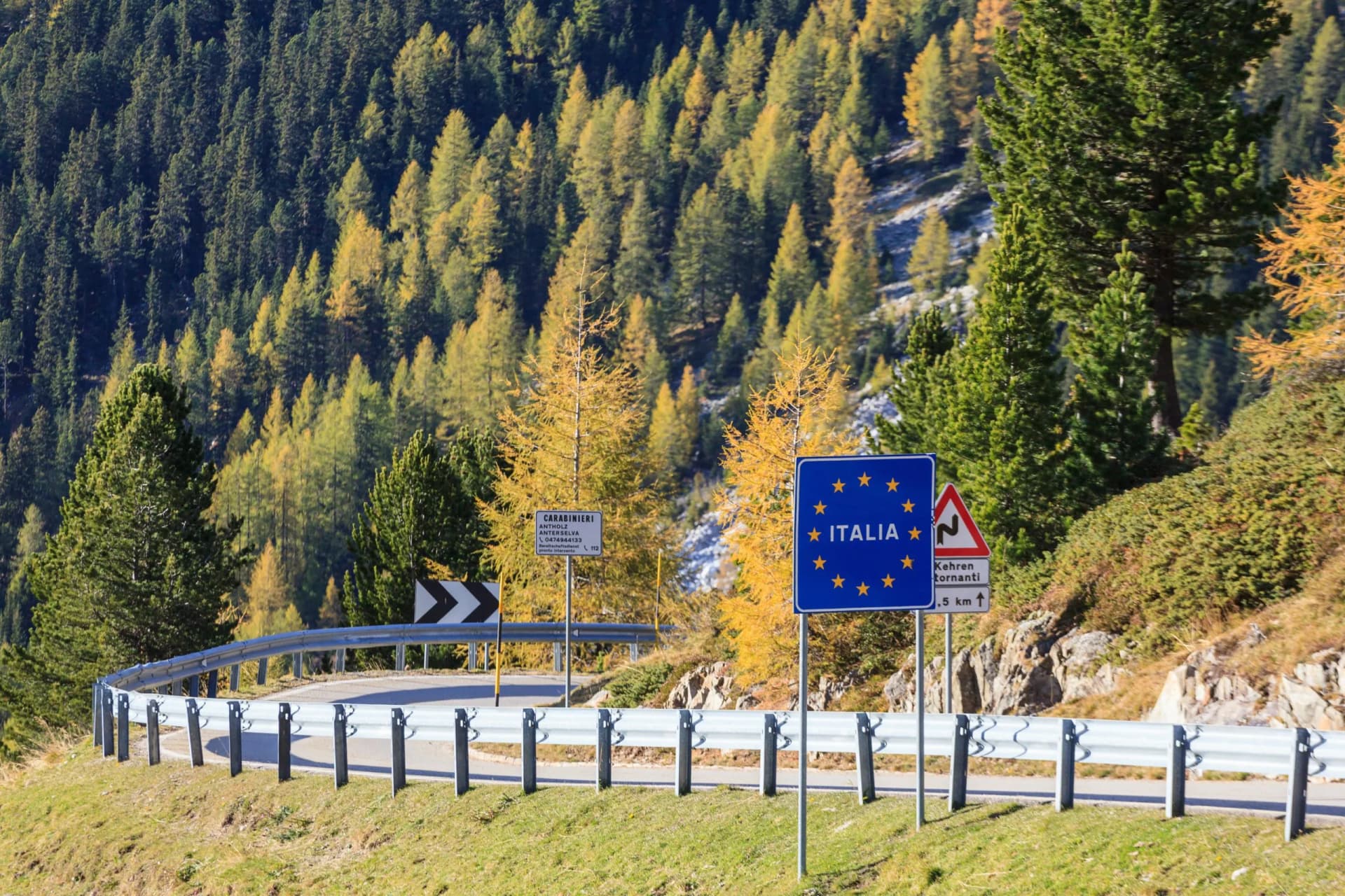 Road sign indicating entry to Italia next to guardrail on mountain road with autumn trees.