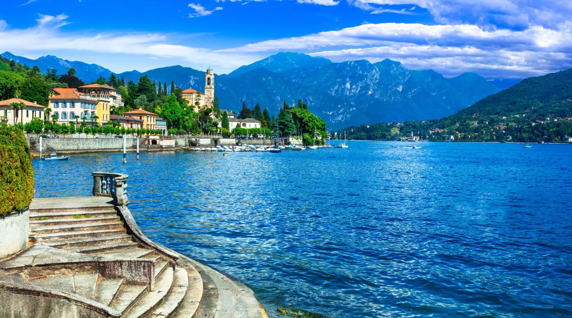 Stone steps leading to deep blue lake water with boats and colorful lakeside town backed by mountains.