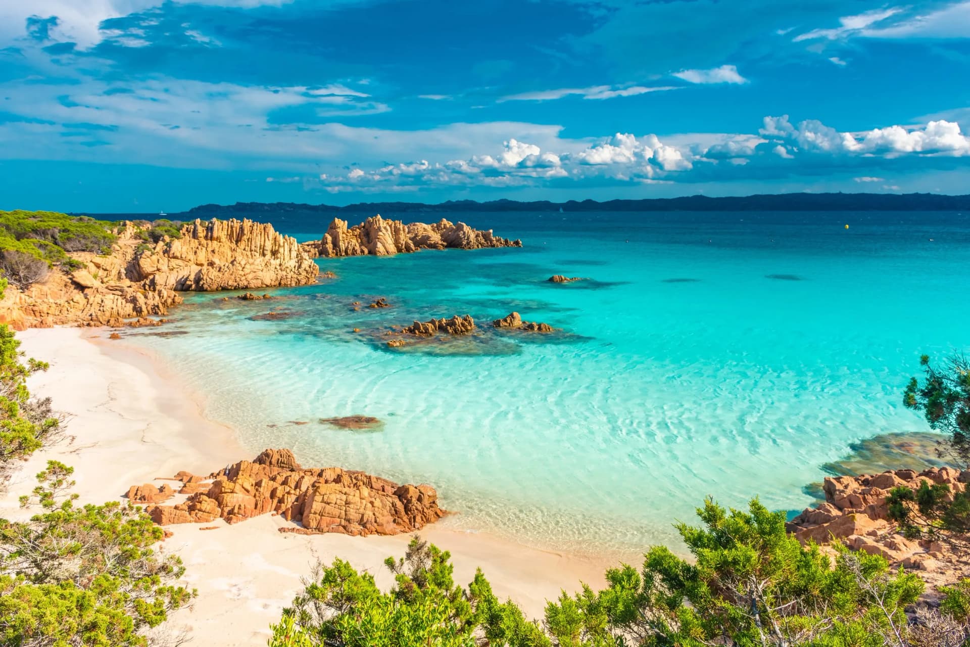 Secluded white sand beach with turquoise water, orange rocks, and green scrub in Sardinia.