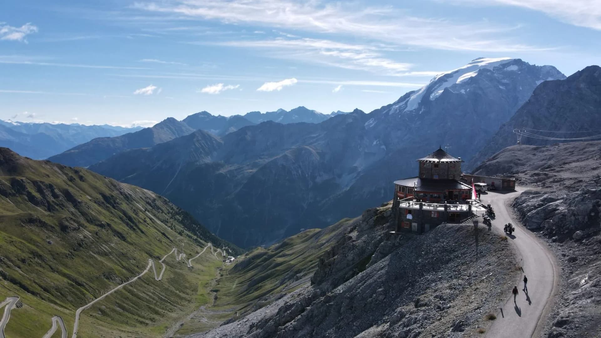 Motorcyclists and hikers near restaurant on Stelvio Pass with winding road and snowy mountains.