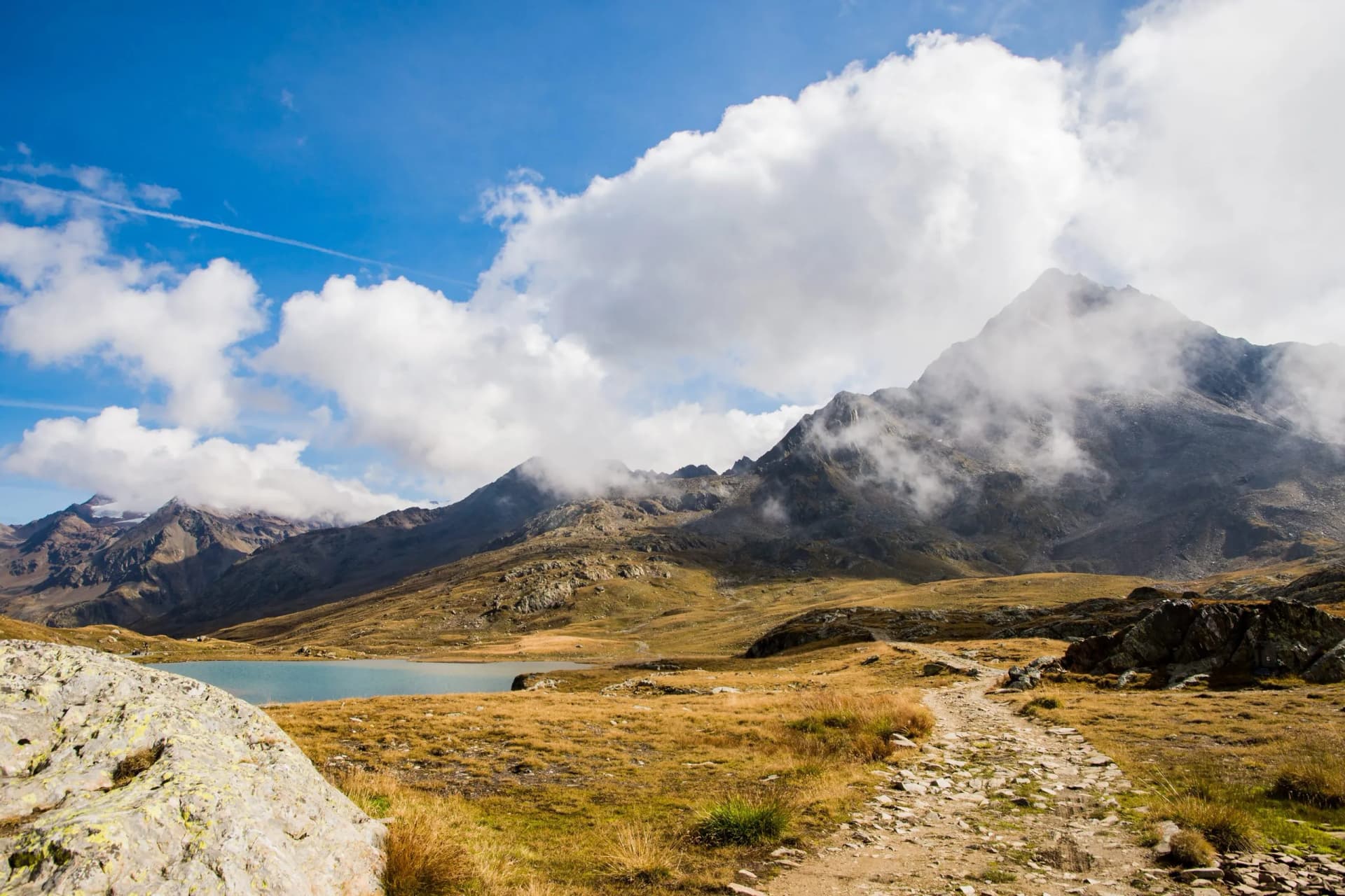 Rocky hiking trail leading toward alpine lake and rugged mountains under cloudy blue sky
