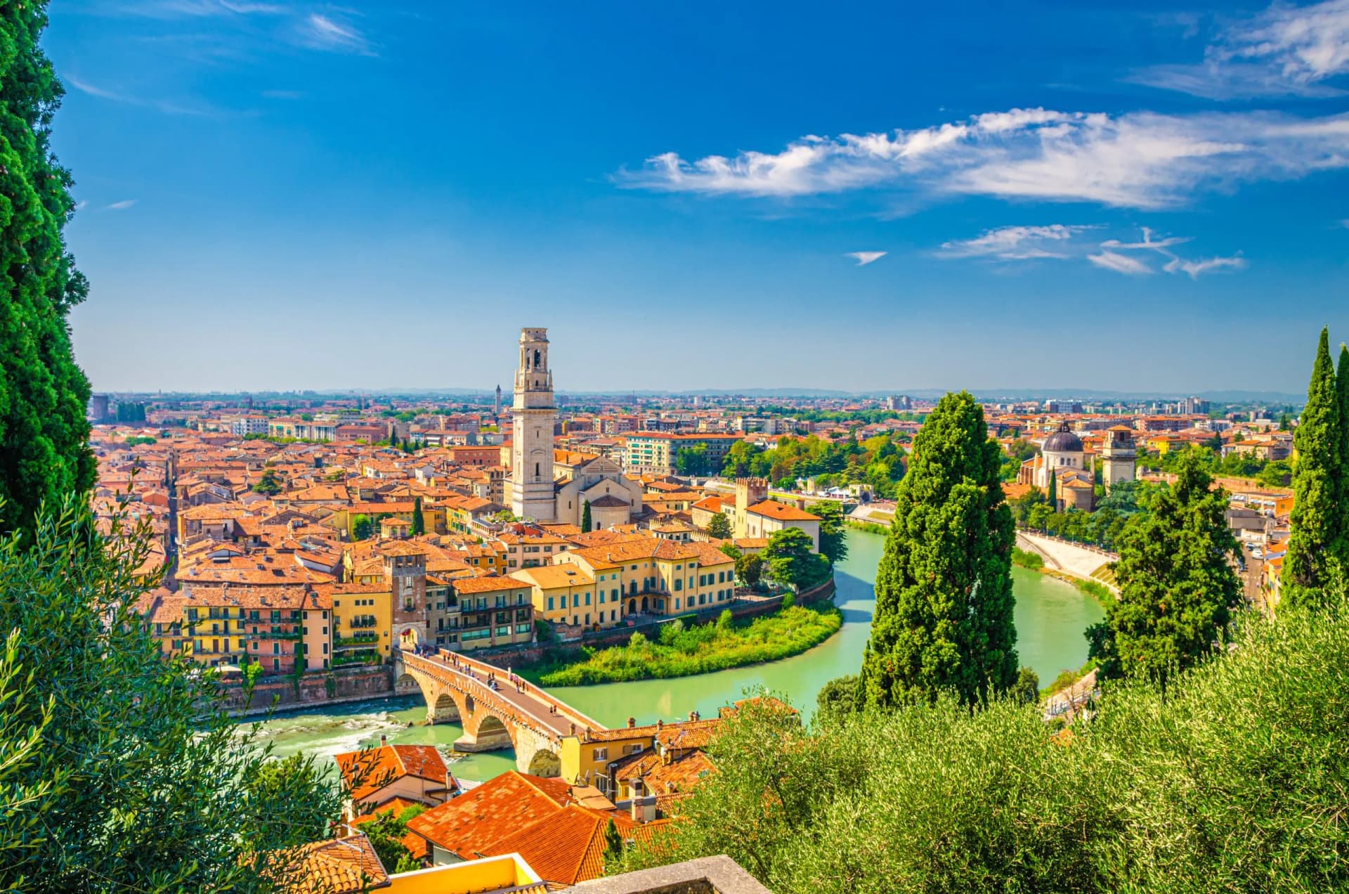 Verona cityscape with terracotta roofs, river, bridge, and tall bell tower under blue sky.