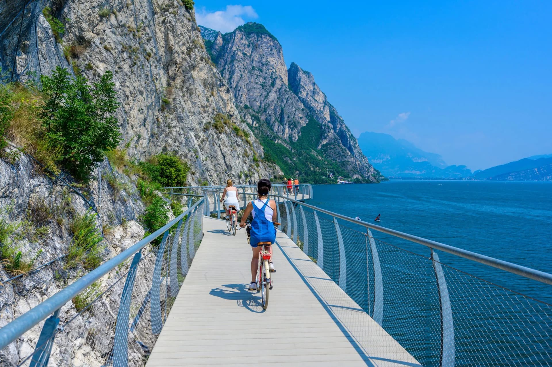 Cyclists on a suspended boardwalk beside a steep cliff overlooking deep blue water, Garda.