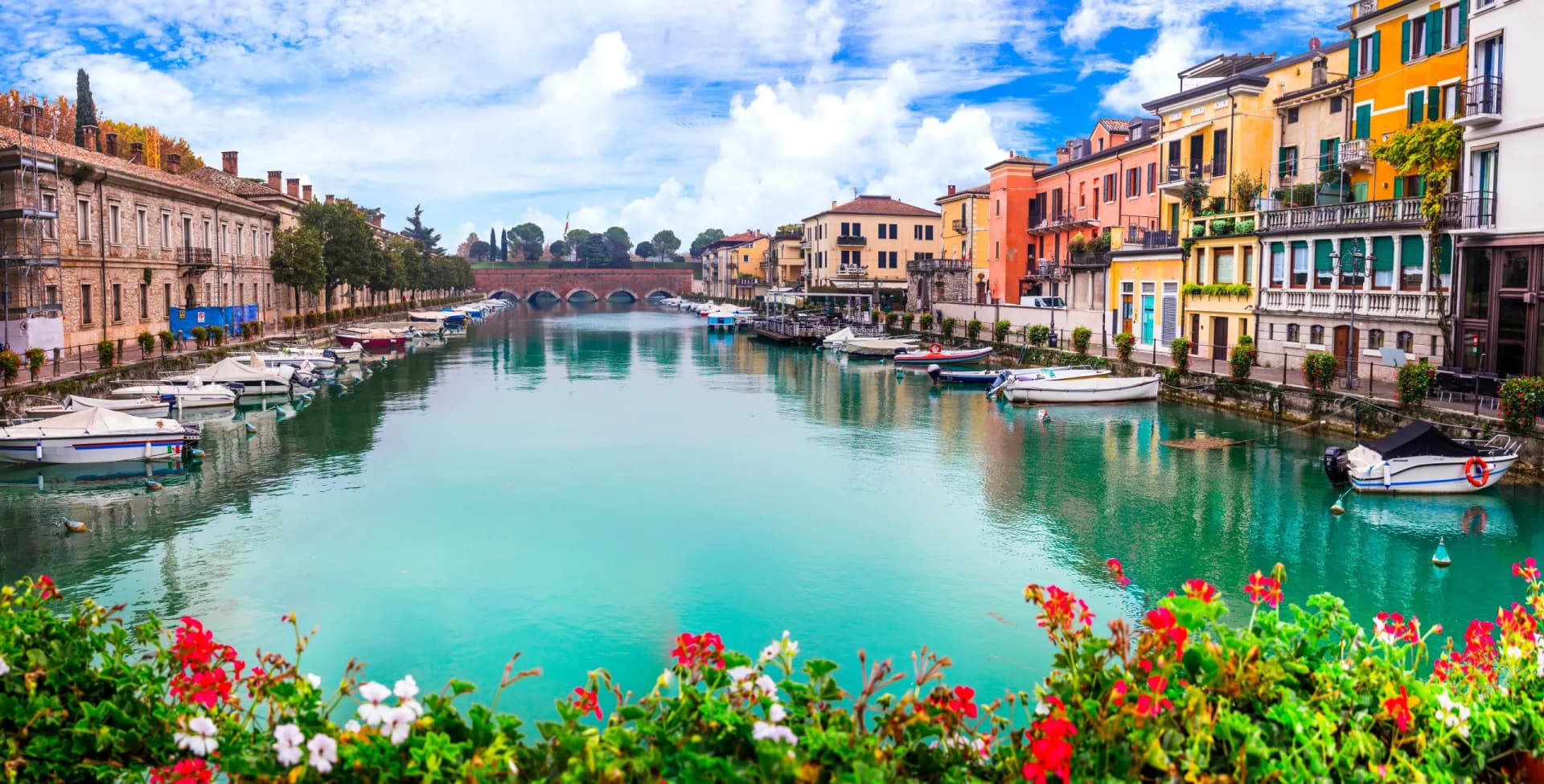 Boats docked on a canal with colorful buildings and a stone bridge in Peschiera.