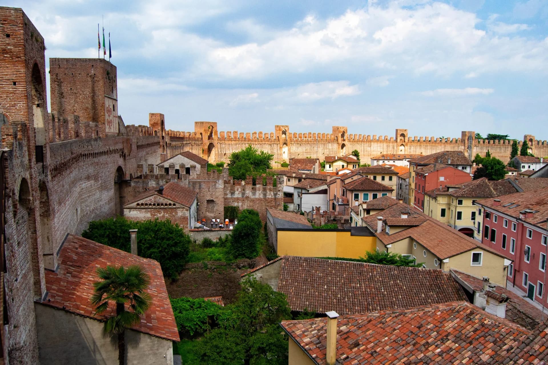 View over terracotta roofs toward the medieval brick walls and towers of Montagnana under a cloudy sky.