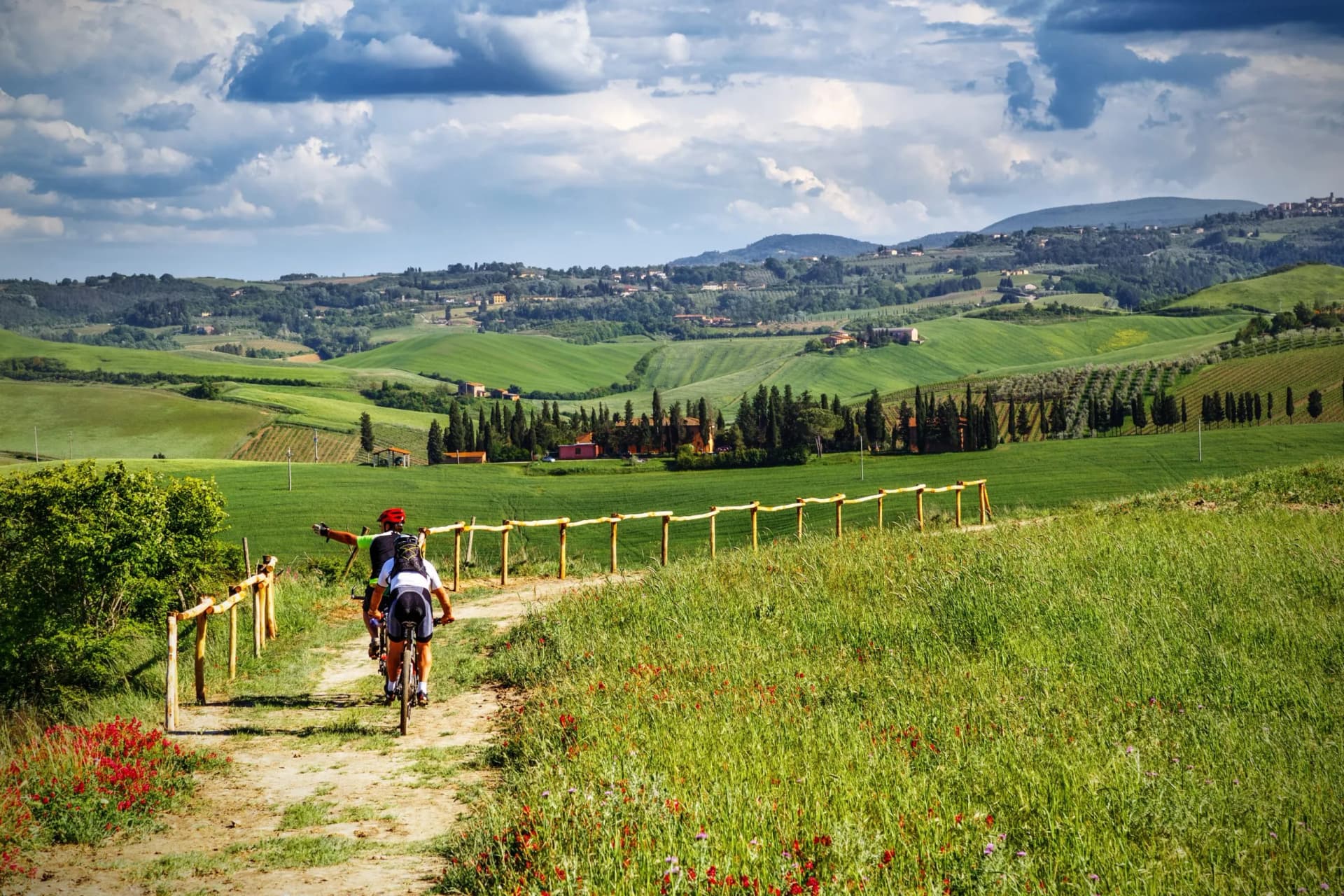 Cyclists on dirt path through rolling green hills and cypress trees in Tuscany.