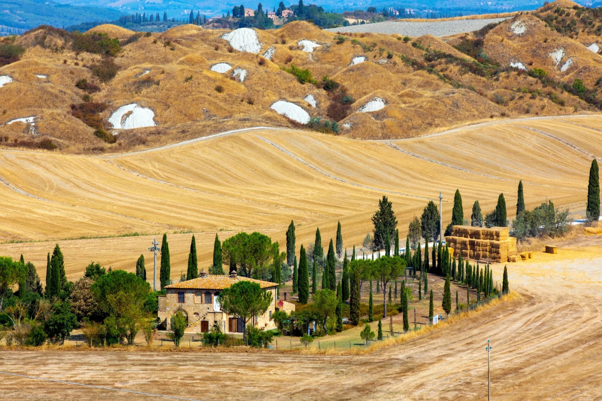 Rolling golden hills of Crete Senesi with a farmhouse, cypress trees, and hay bales.