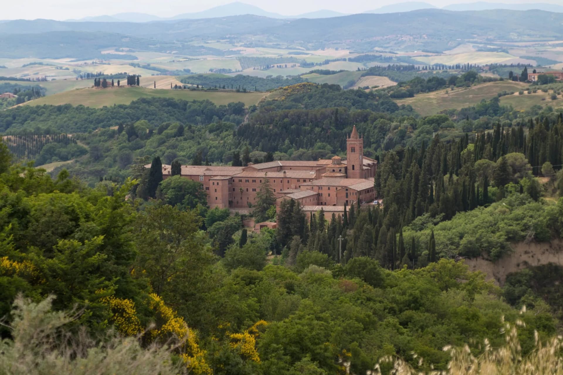 Monastery building nestled in lush green hills with rolling Tuscan landscape background.