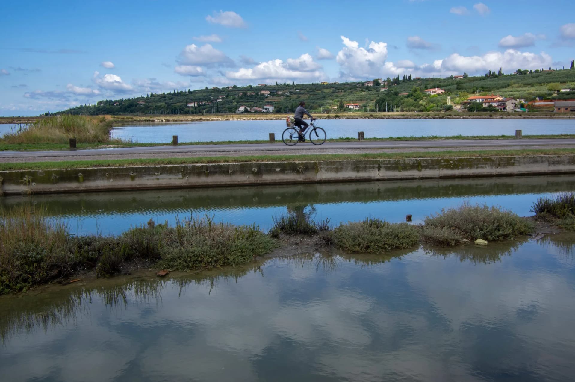 Cyclist riding along road next to water pools with hillside houses in background