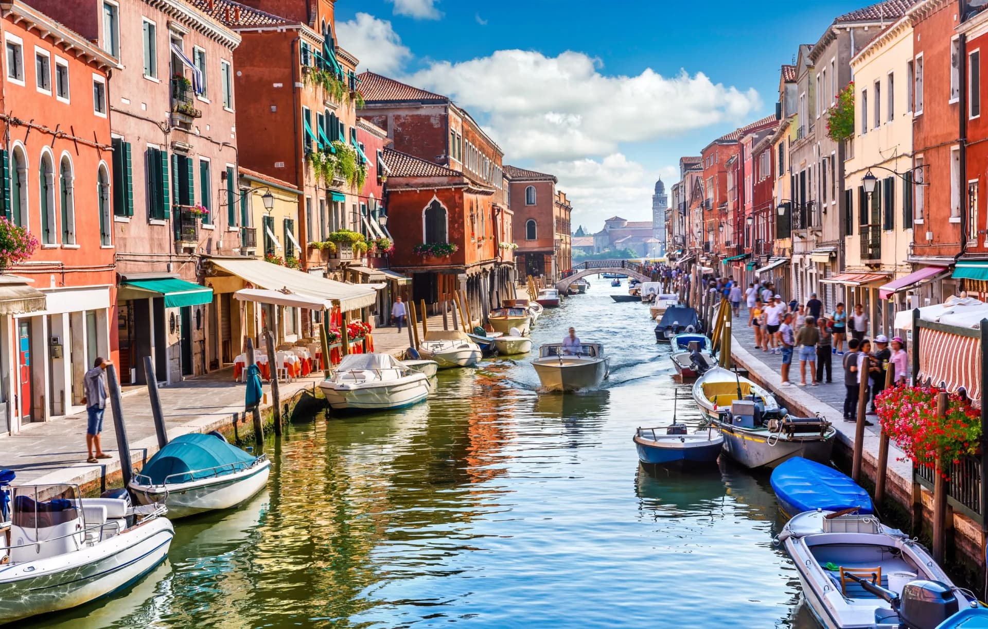 Boats on a canal lined with colorful buildings and pedestrians in Venice, Italy.