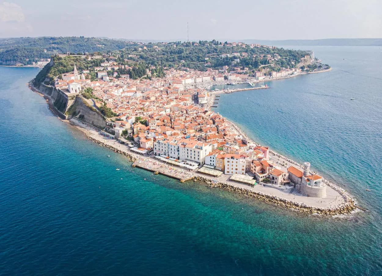Aerial view of Slovenian coast and Piran peninsula with red-roofed town buildings and clear blue sea.