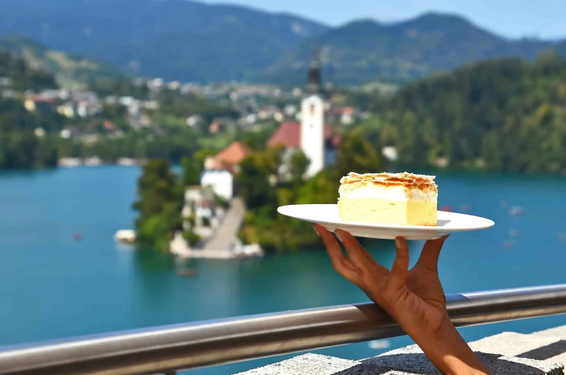 Cream cake slice held over Lake Bled with island church and mountains, Slovenia