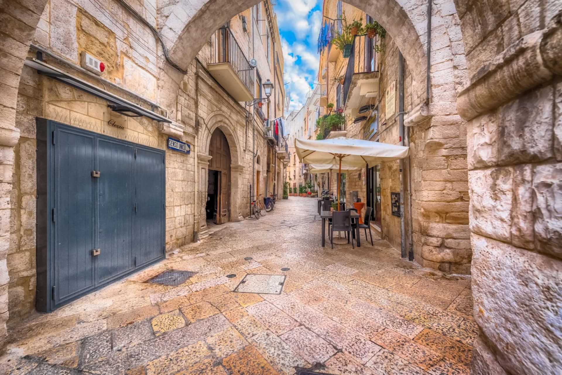 Narrow stone alleyway in Bari with an outdoor cafe under umbrellas and blue sky visible.