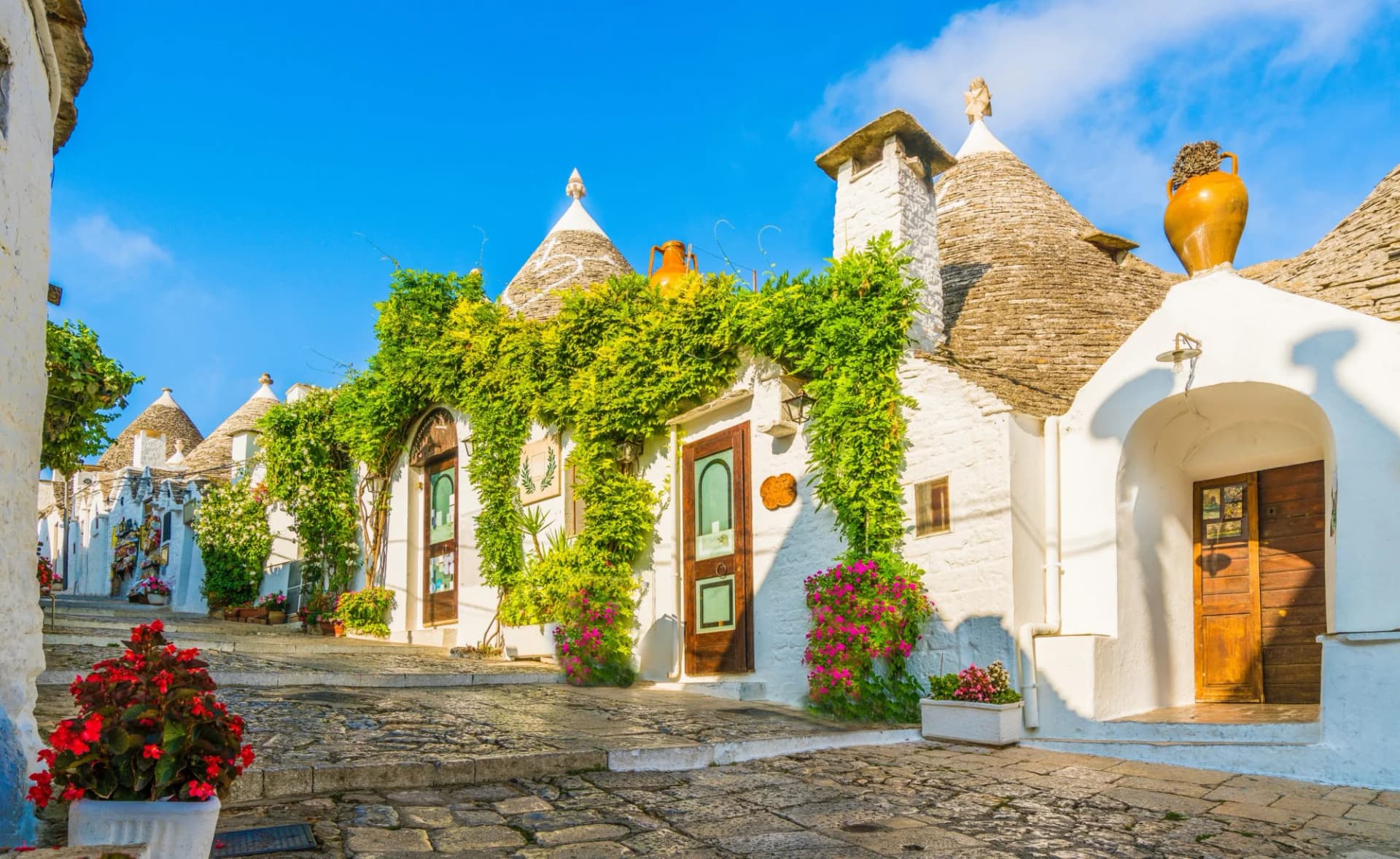 Trulli houses with conical stone roofs on a cobblestone street under a bright blue sky.