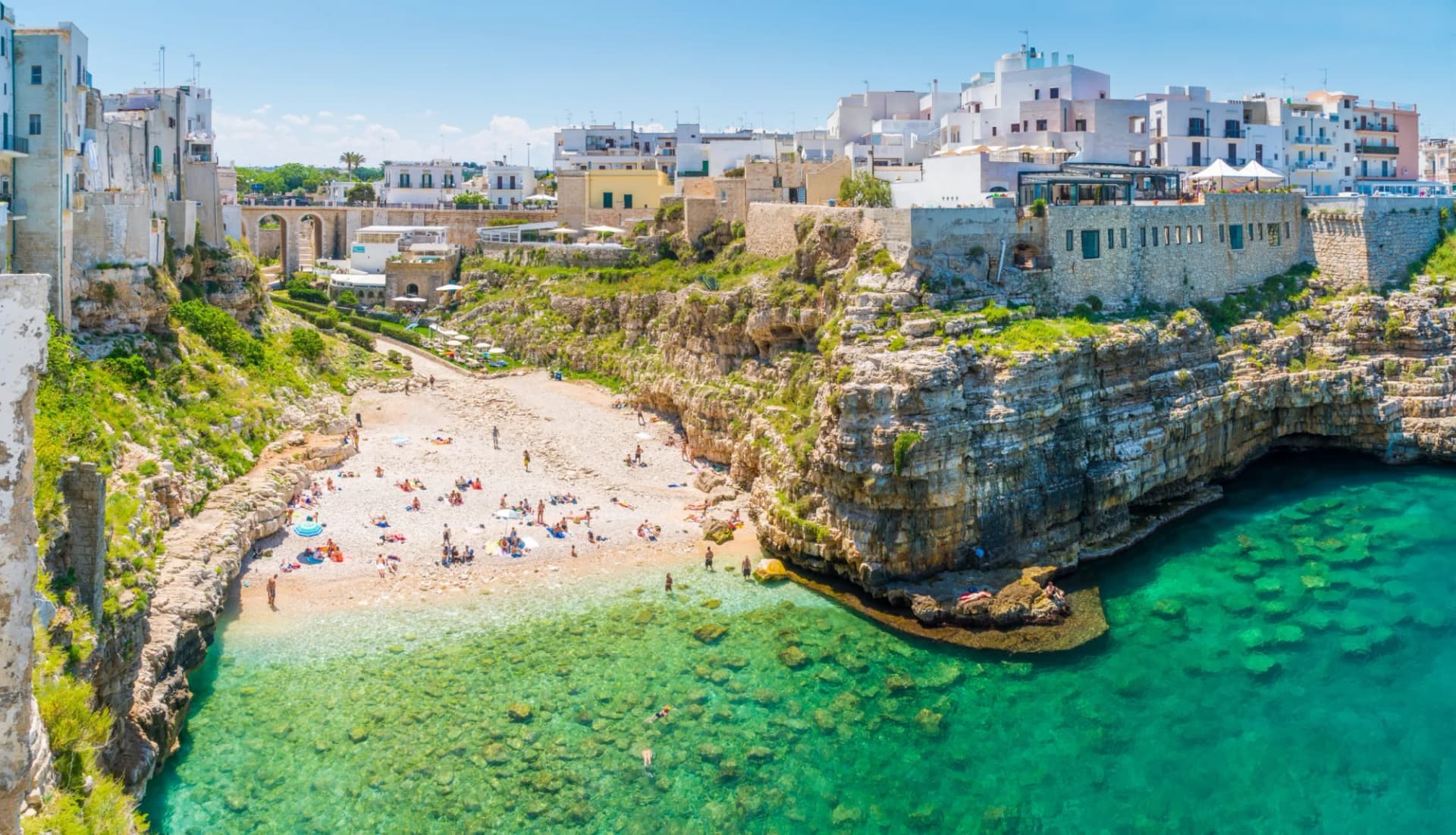 Beachgoers sunbathing by turquoise sea below white buildings on cliffside in Bari region.