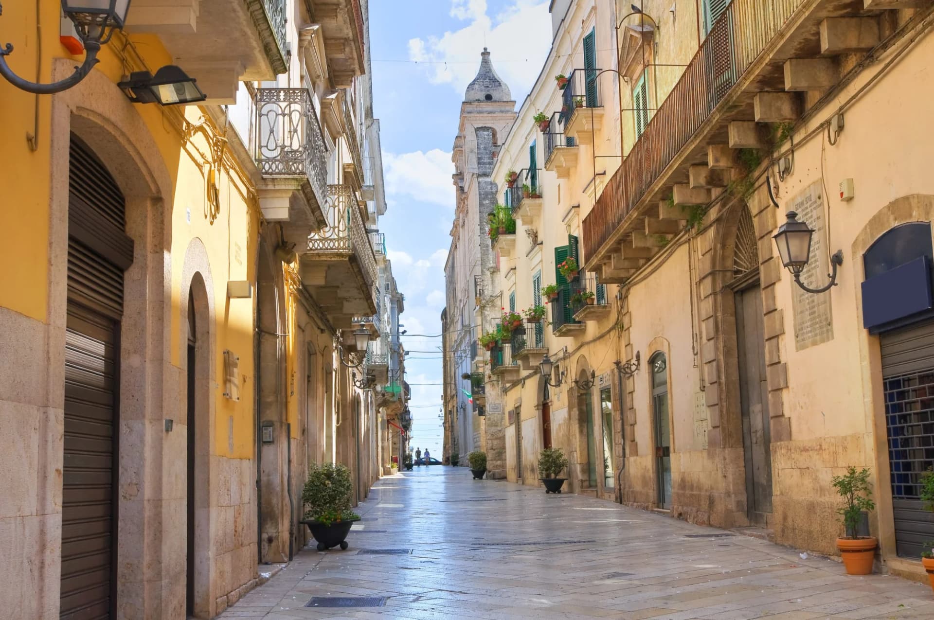 Narrow stone street in Altamura leading to a church tower under a blue sky.