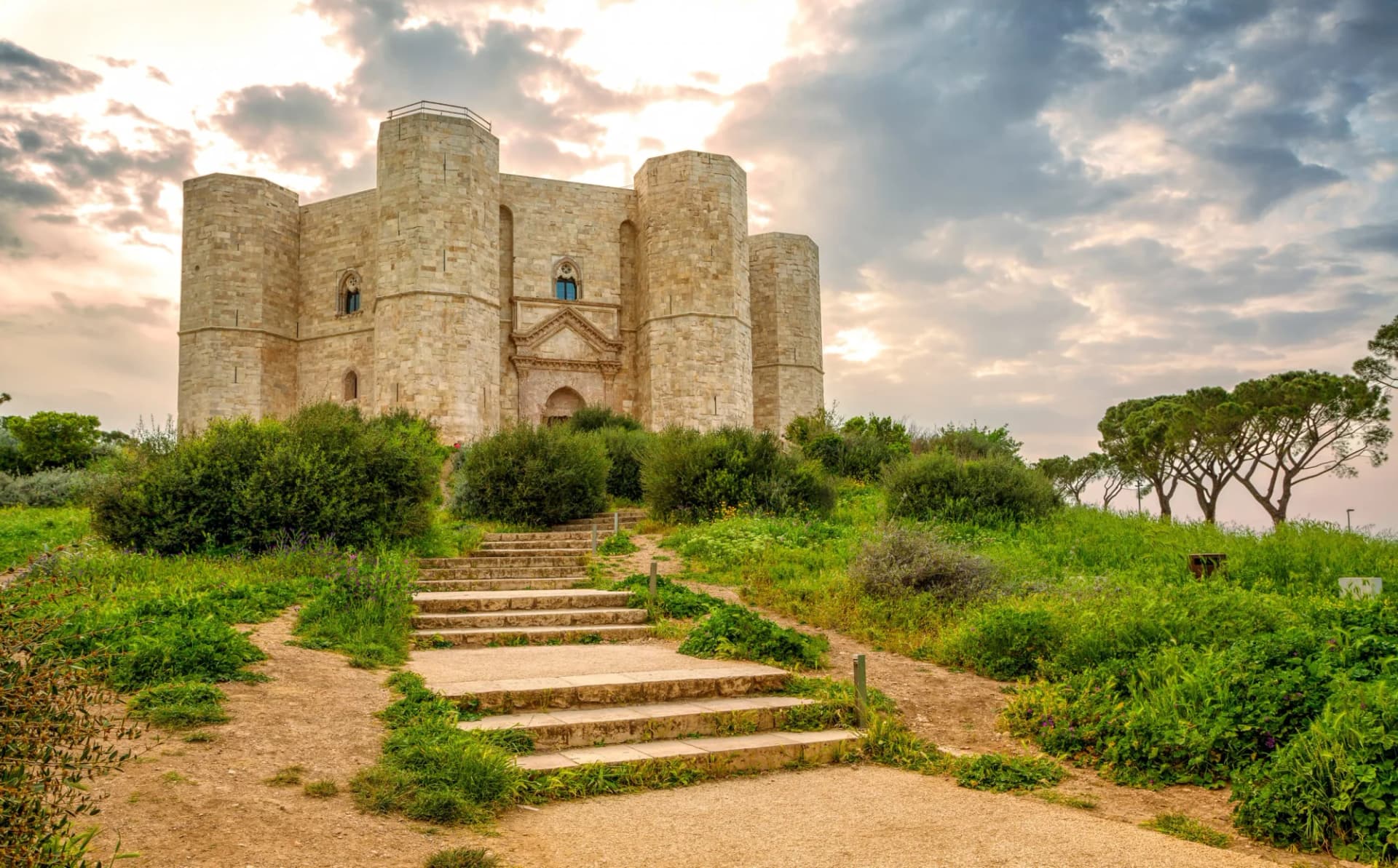 Castel del Monte stone fortress approached by stone steps through green hillside under cloudy sky.