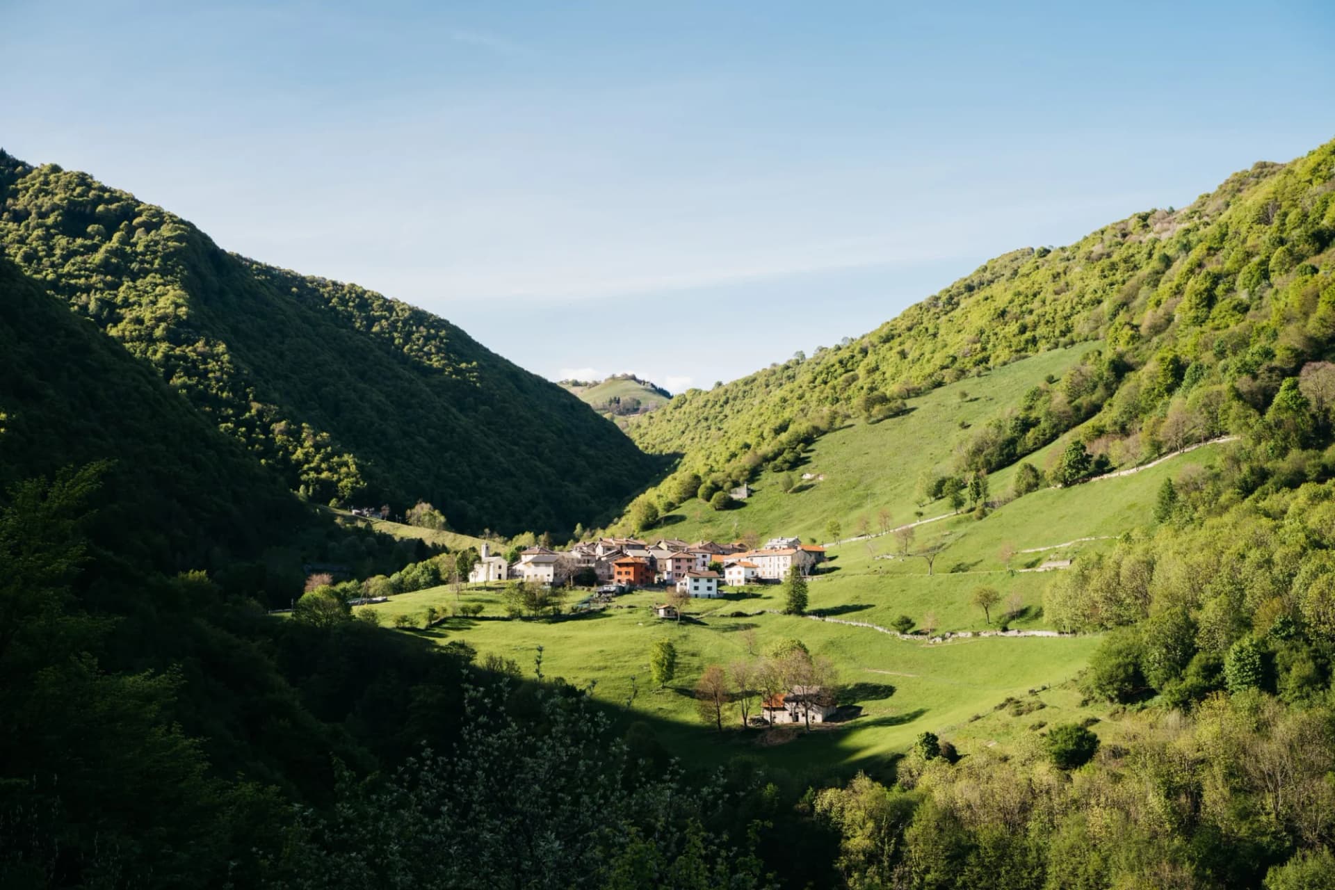 Small village nestled in a green valley between steep, forested mountains under a clear blue sky.