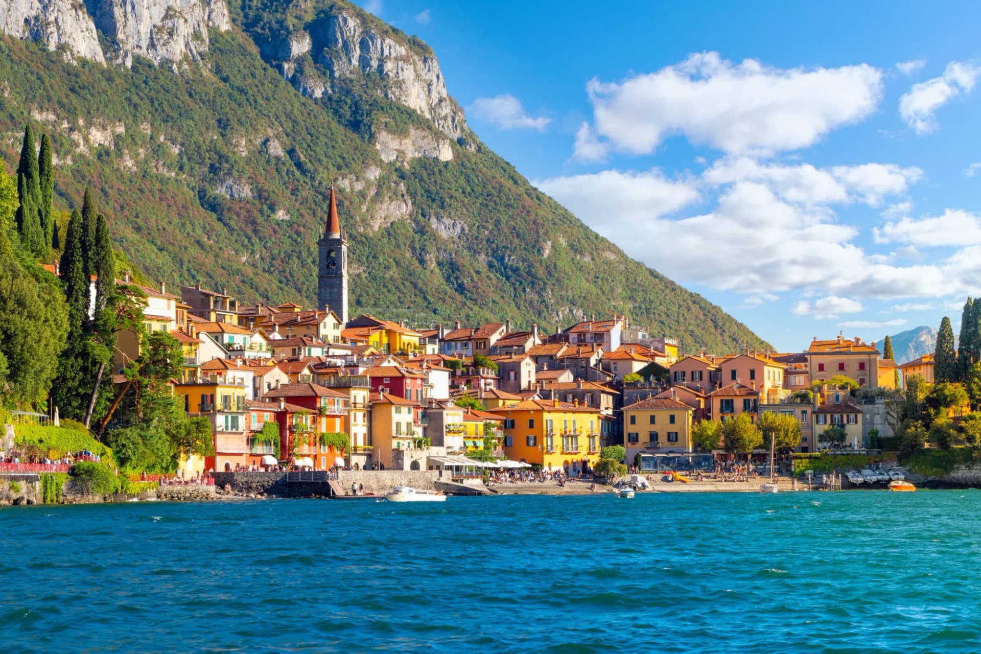 Colorful village on Lake Como with boats, steep green mountains, and blue sky.