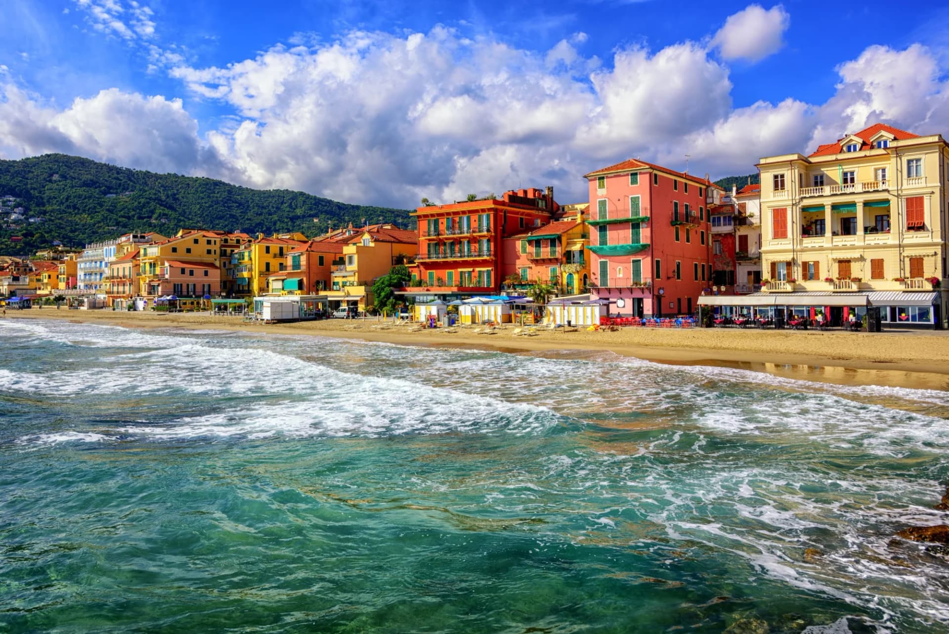 Waves breaking on beach in front of colorful buildings backed by green hills, San Remo.