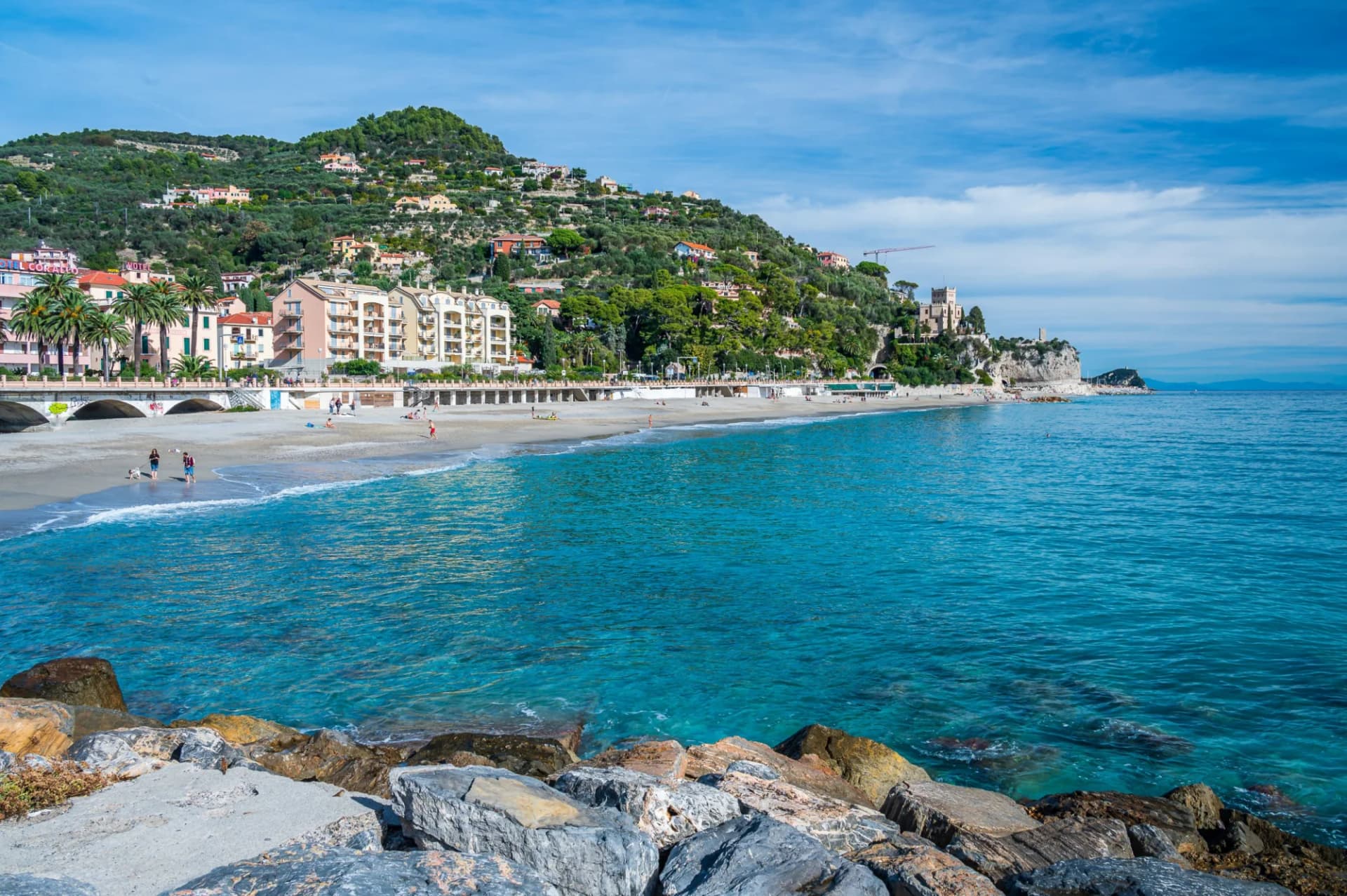 Beach with turquoise water, buildings on a green hill, and a castle on a cliff in Finale Ligure.