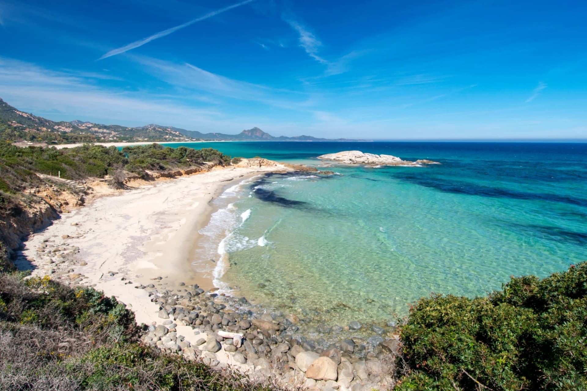Sandy cove with turquoise water, rocks, and green coastal vegetation under a blue sky.