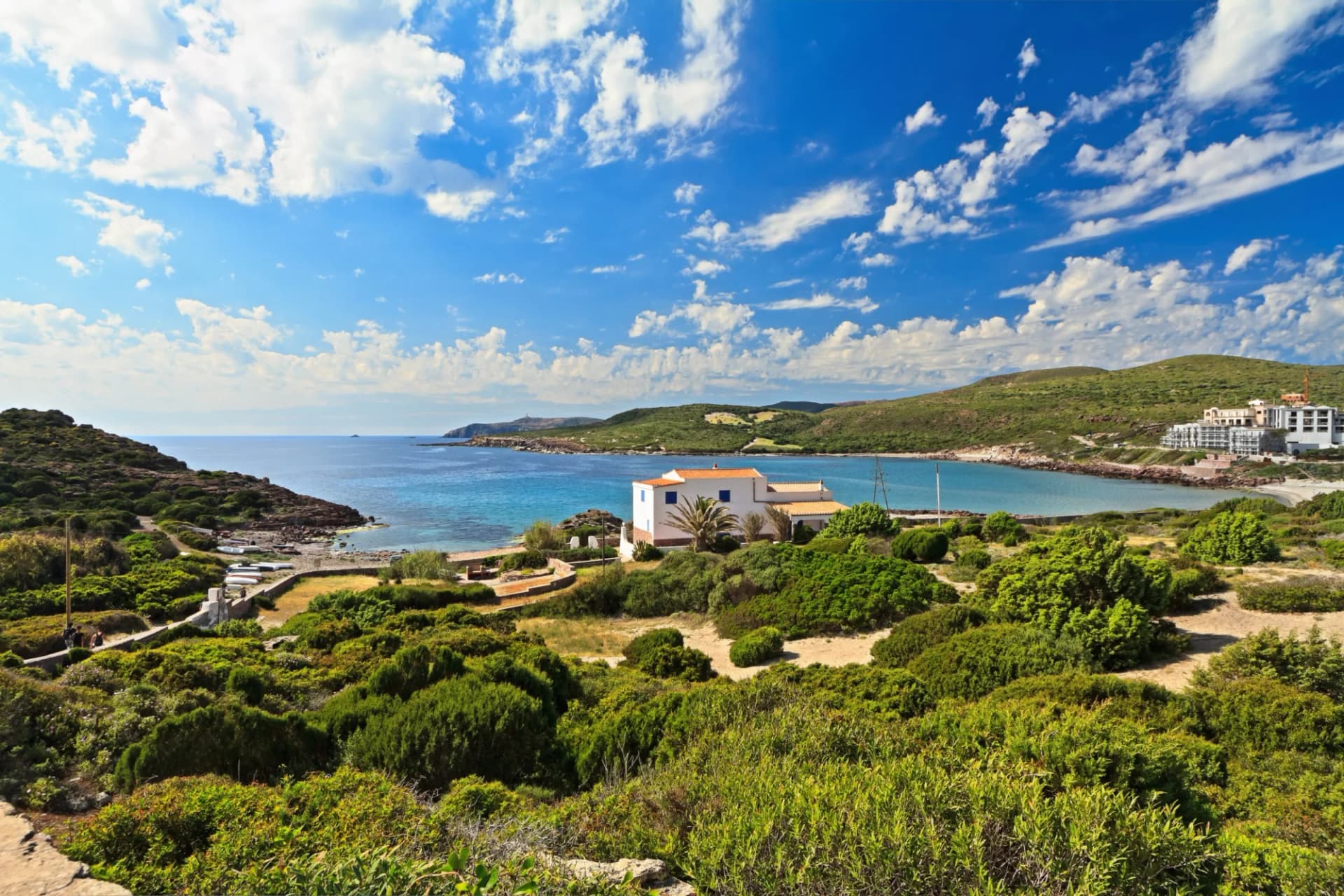 Coastal bay with turquoise water, white house, and green hills under a blue sky with clouds.