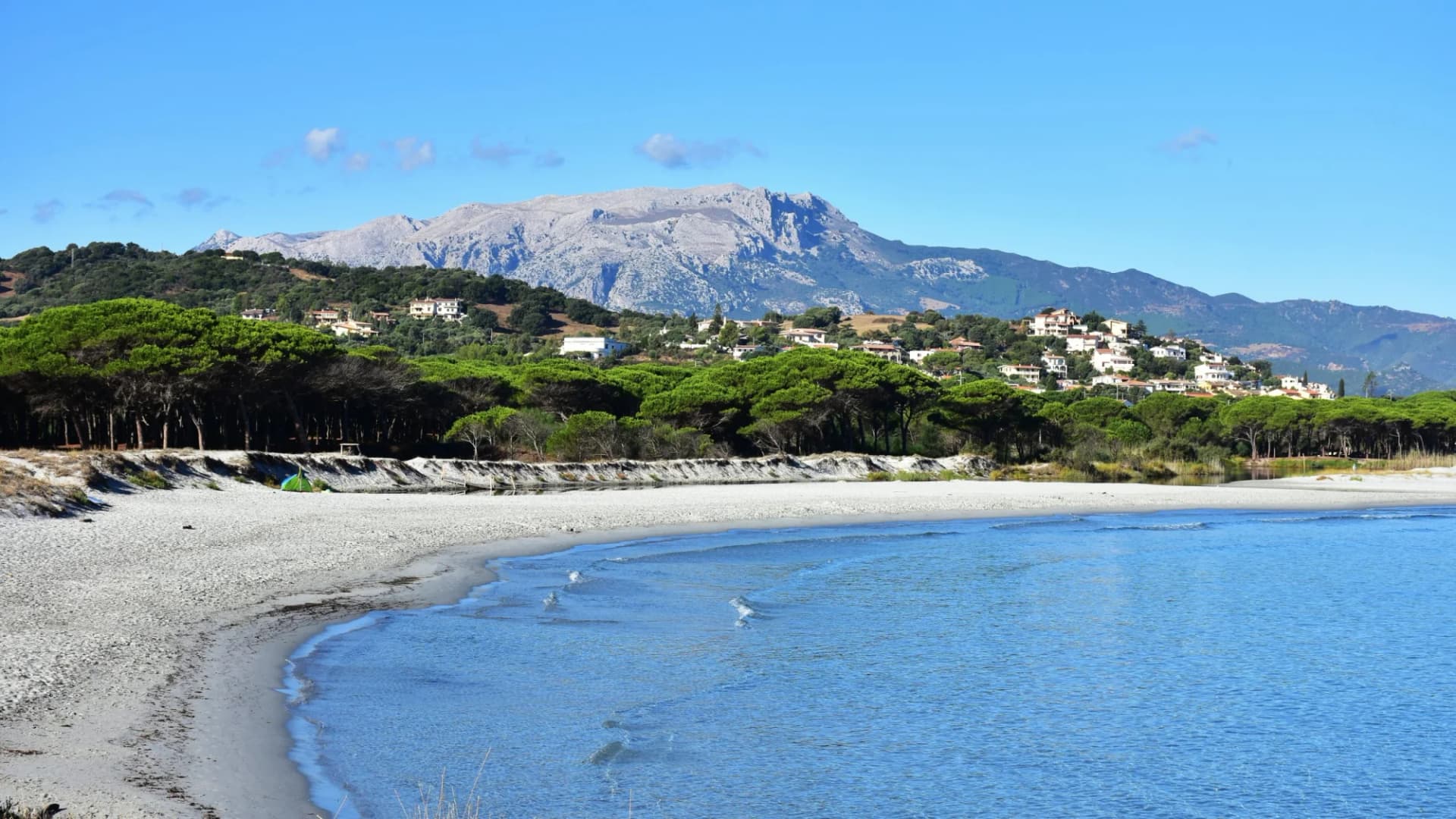 White sand beach meeting turquoise water with pine trees and mountain village in Sardinia