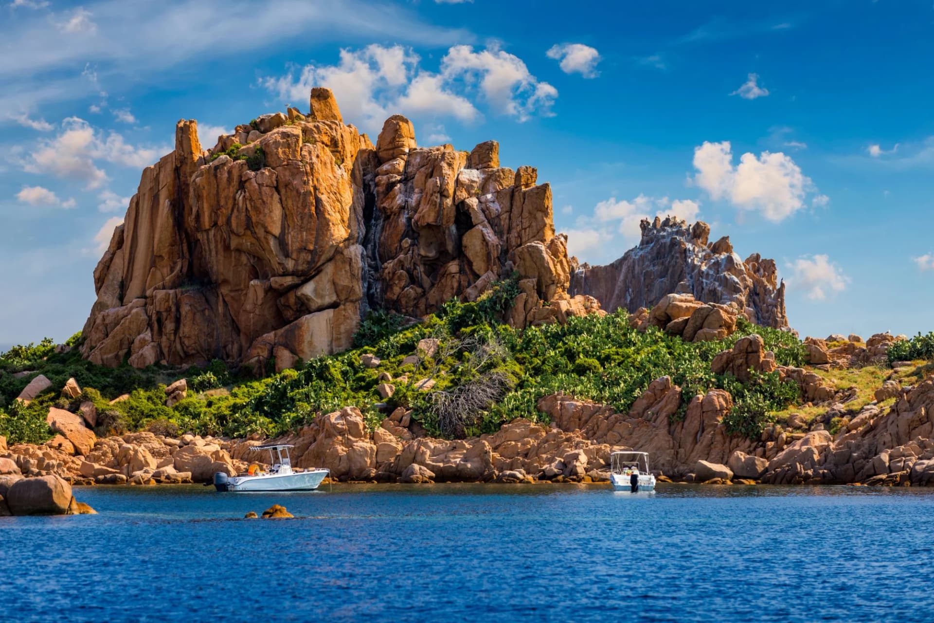 Two small boats anchored near rugged, sunlit coastal rocks with green scrub under a blue sky.