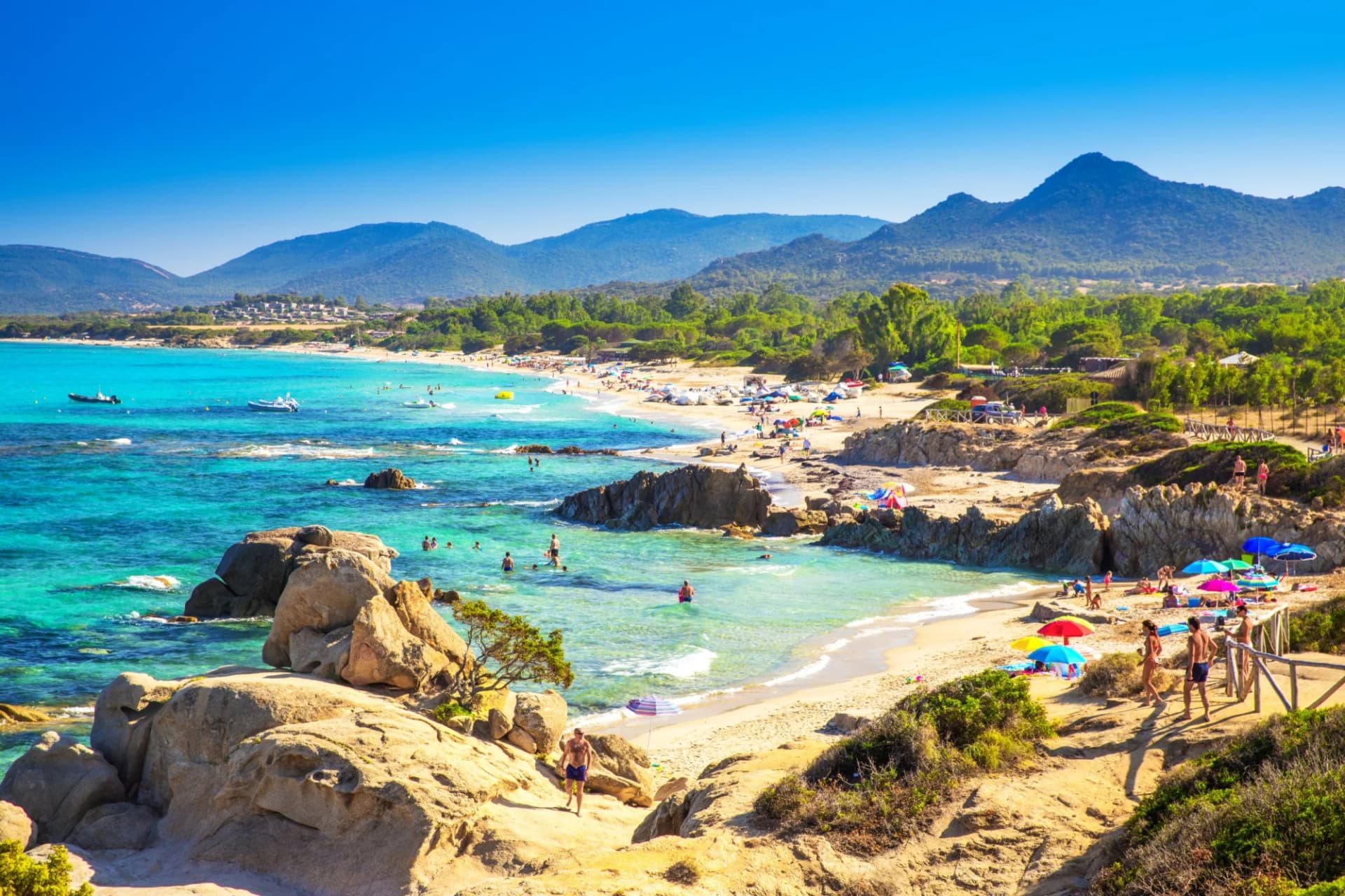 Beachgoers enjoying turquoise water with boats, rocks, and green mountains in the background.