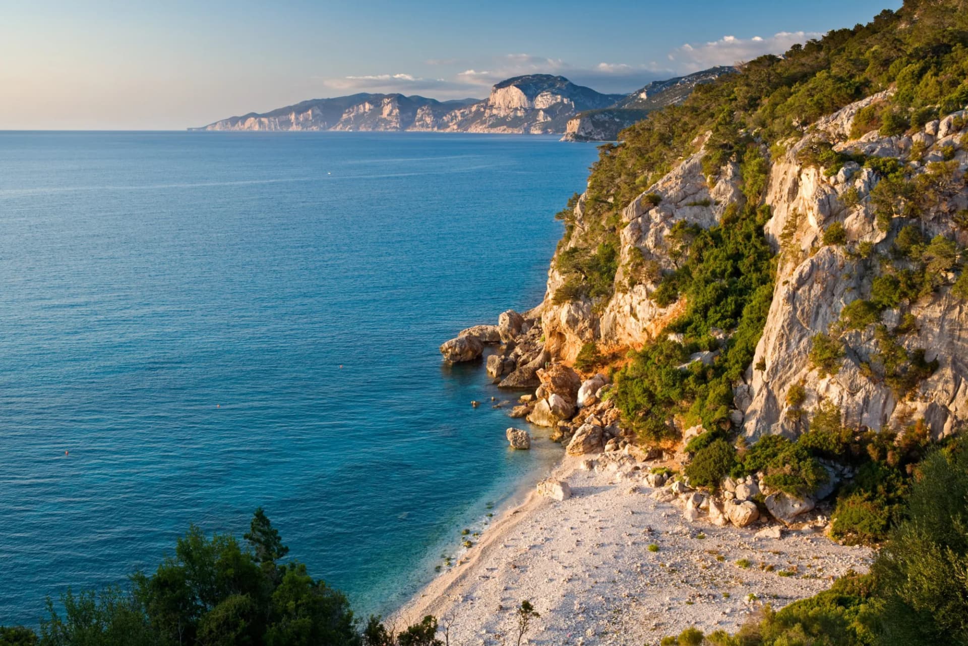 Rocky coastline with scrub brush meeting turquoise sea and distant mountains, Cala Gonone area.