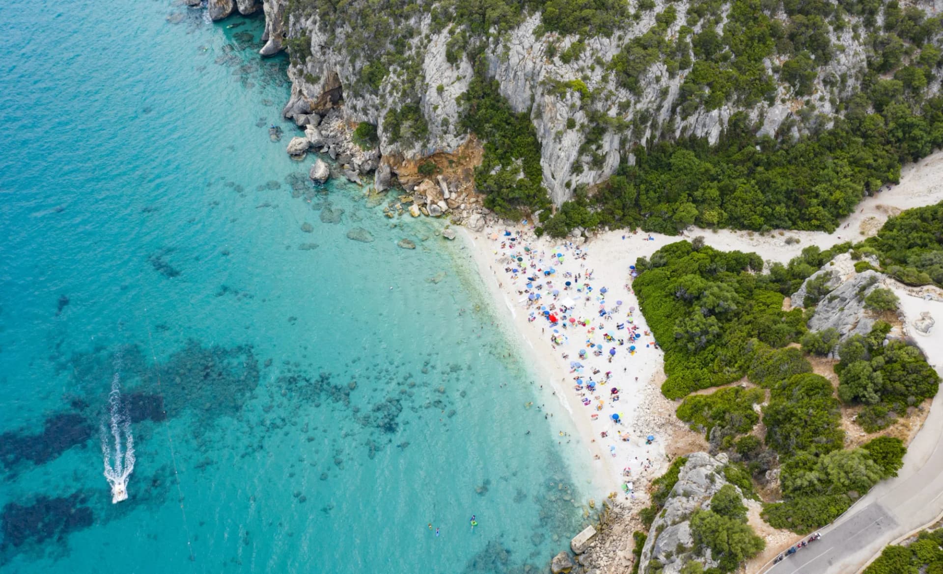 Boat speeding on turquoise water near crowded beach nestled between cliffs in Cala Gonone.