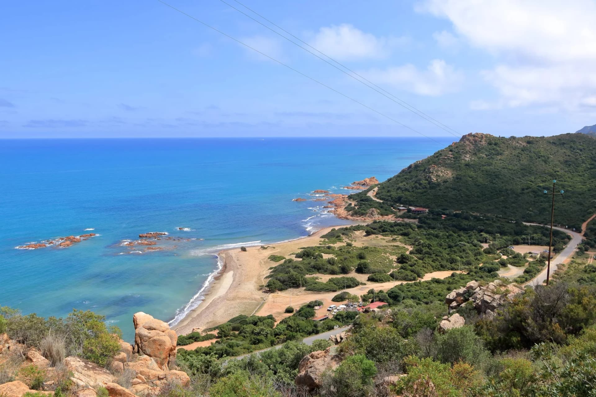 Coastal view of turquoise sea, sandy beach, and green hillsides under a blue sky.