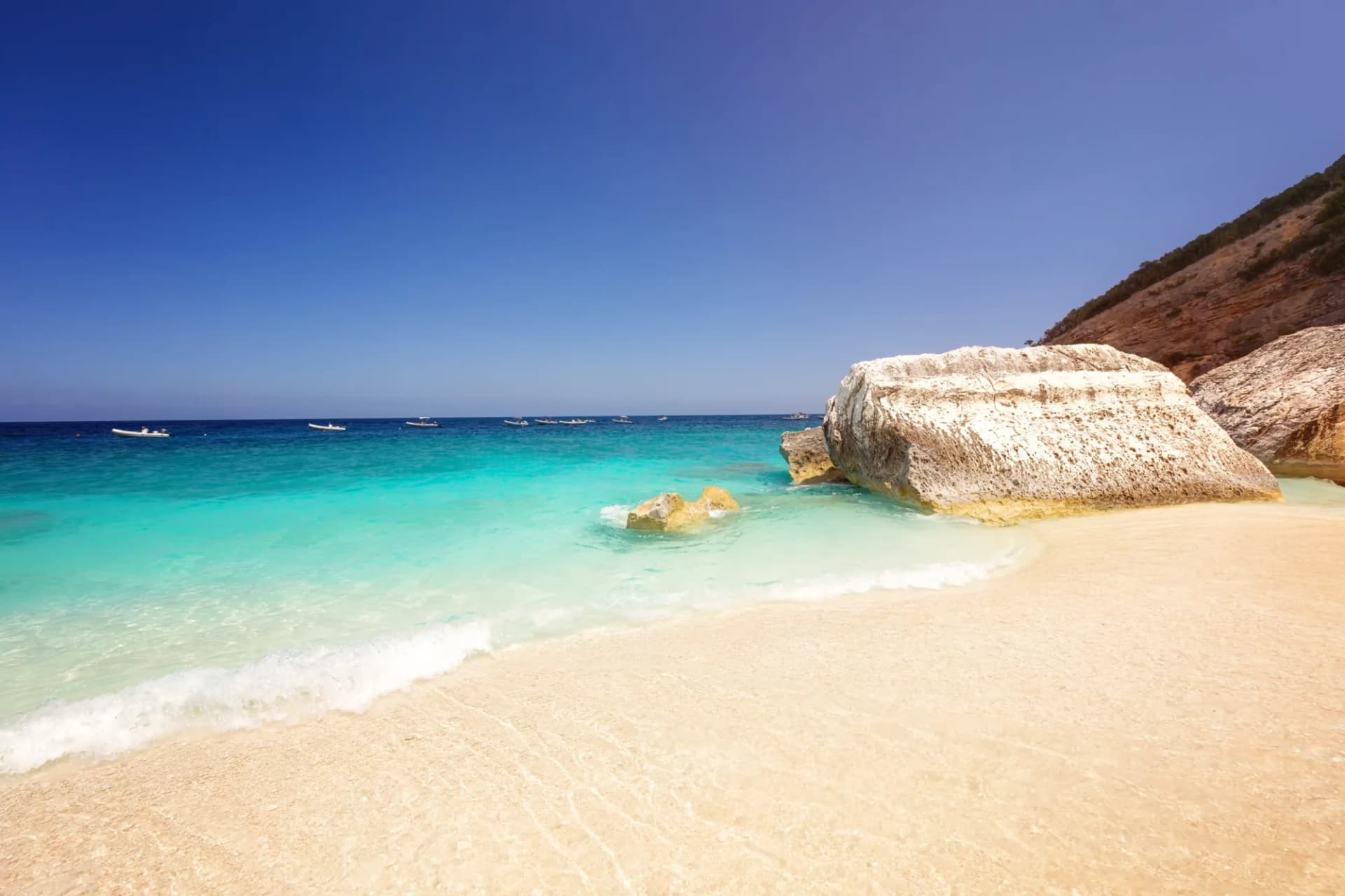 Sandy beach with turquoise water, large white rocks, and boats anchored offshore under a clear blue sky.
