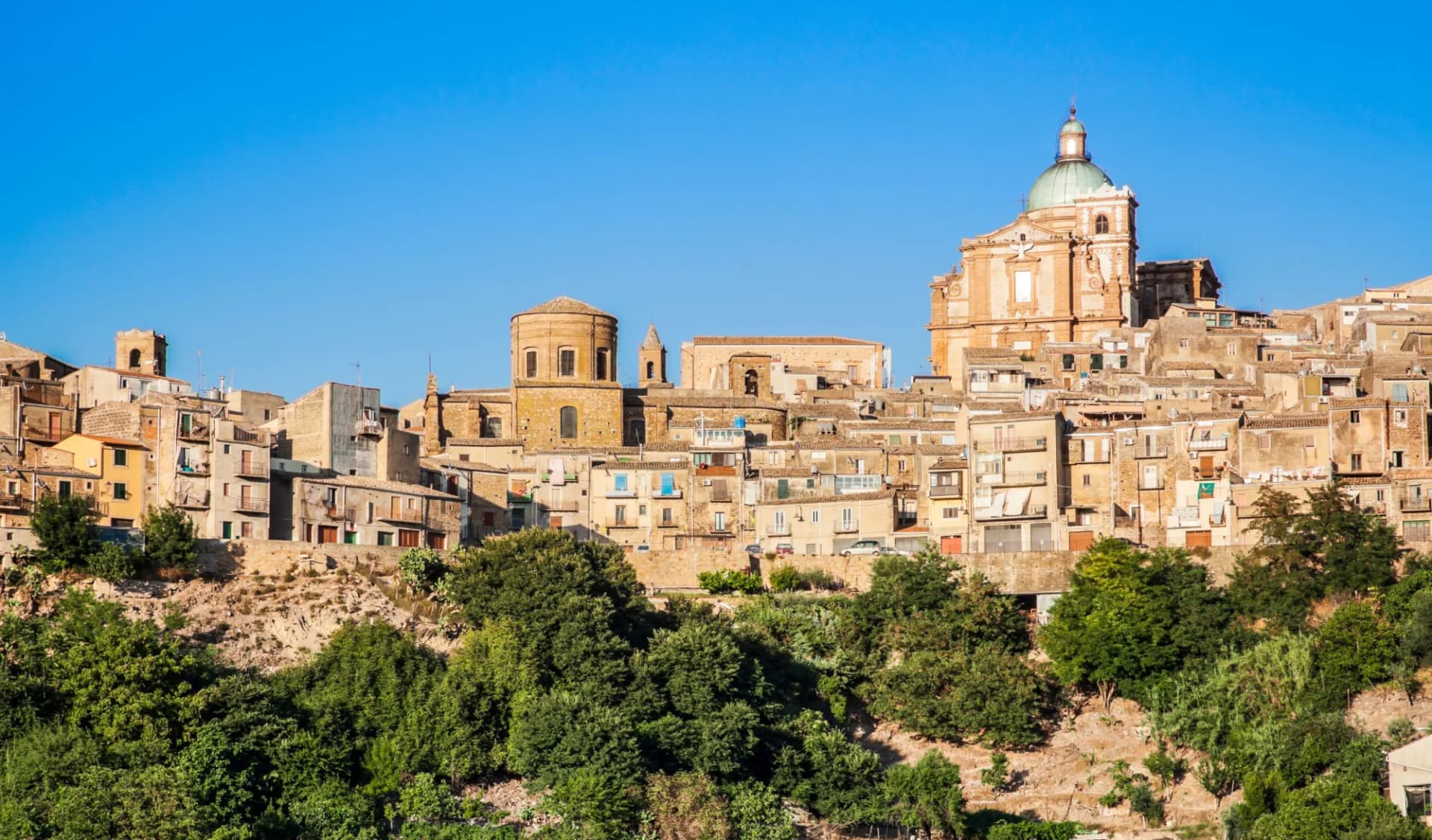 Hillside town with dense historic buildings and prominent church domes under a clear blue sky.