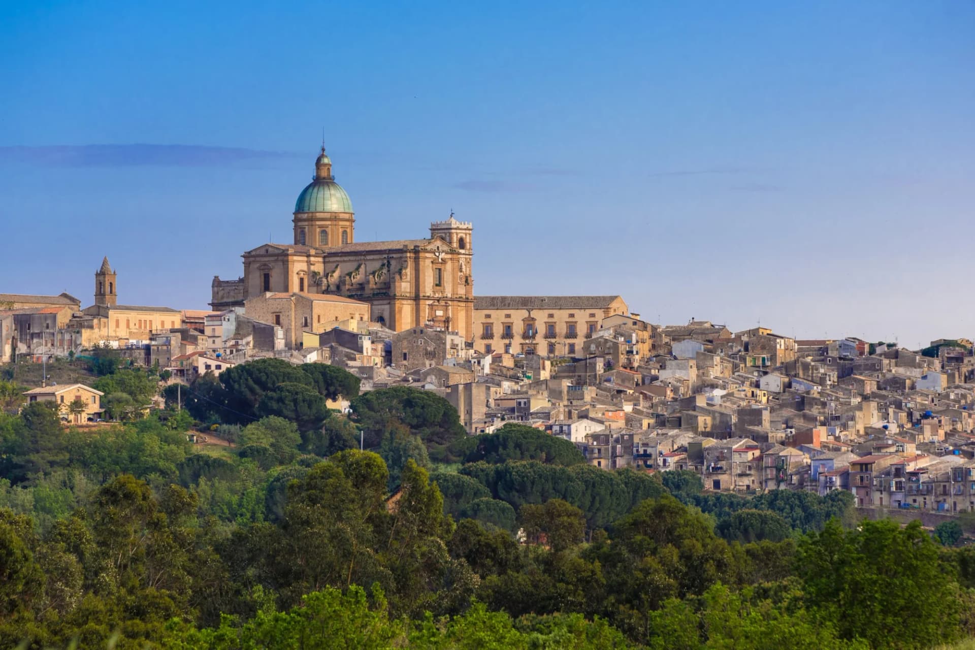 Hillside town with a large domed cathedral above dense green trees under a clear blue sky.