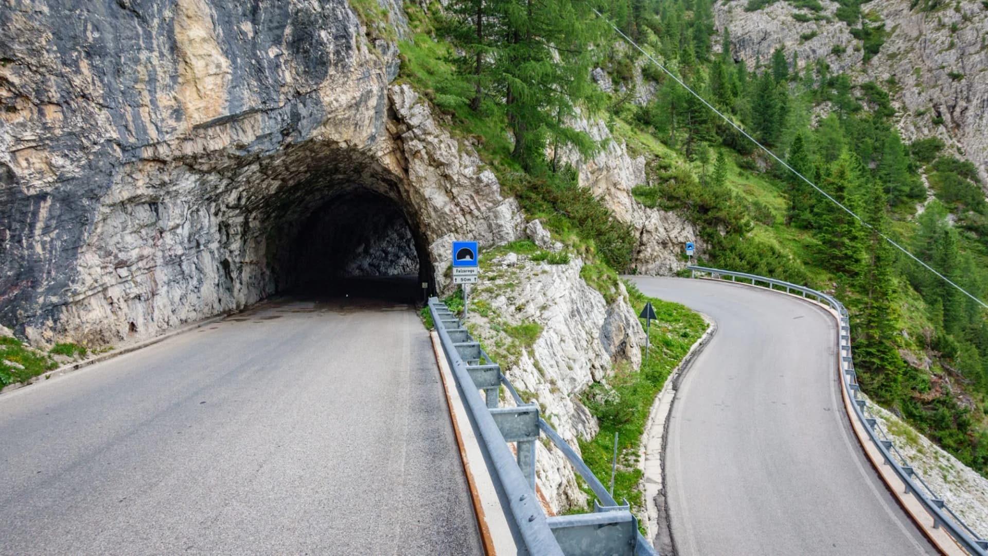 Road curving past Falzarego Pass tunnel entrance carved into rocky, green mountainside.