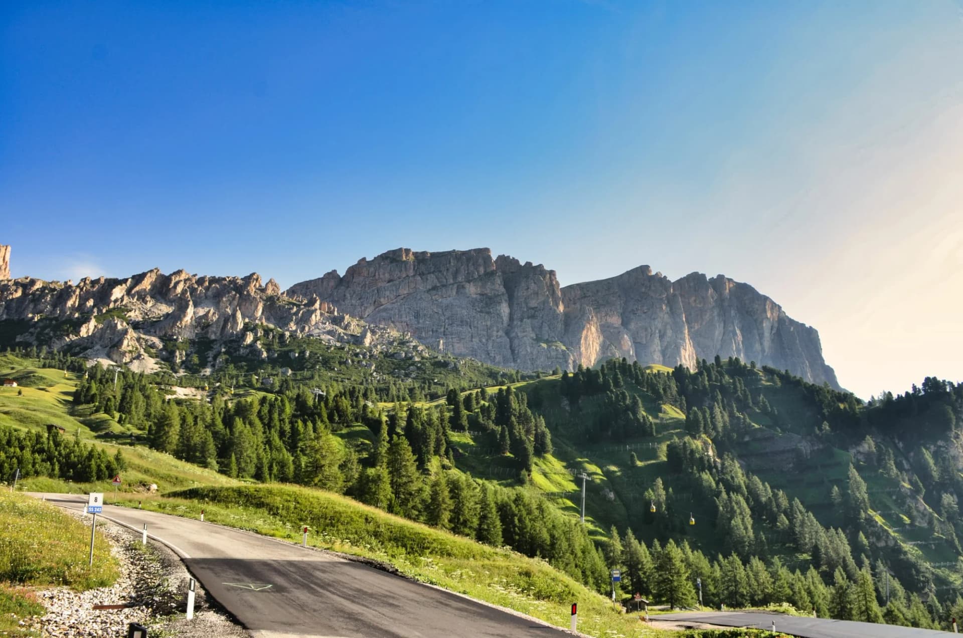 Winding mountain road near Passo Gardena with steep rocky peaks and green forests under a clear blue sky.