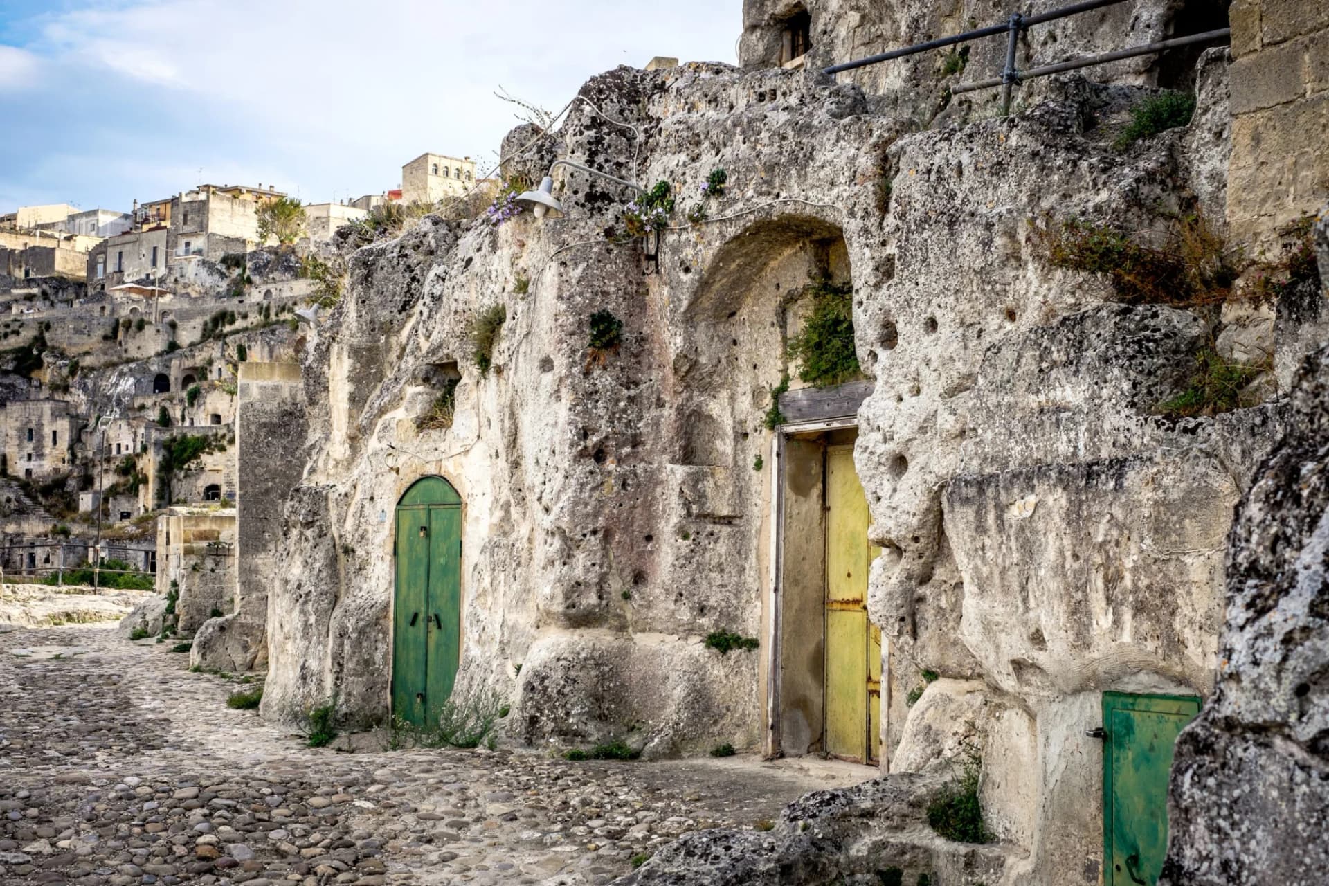 Cave dwellings carved into rock with green and yellow doors on a cobblestone street in Matera.