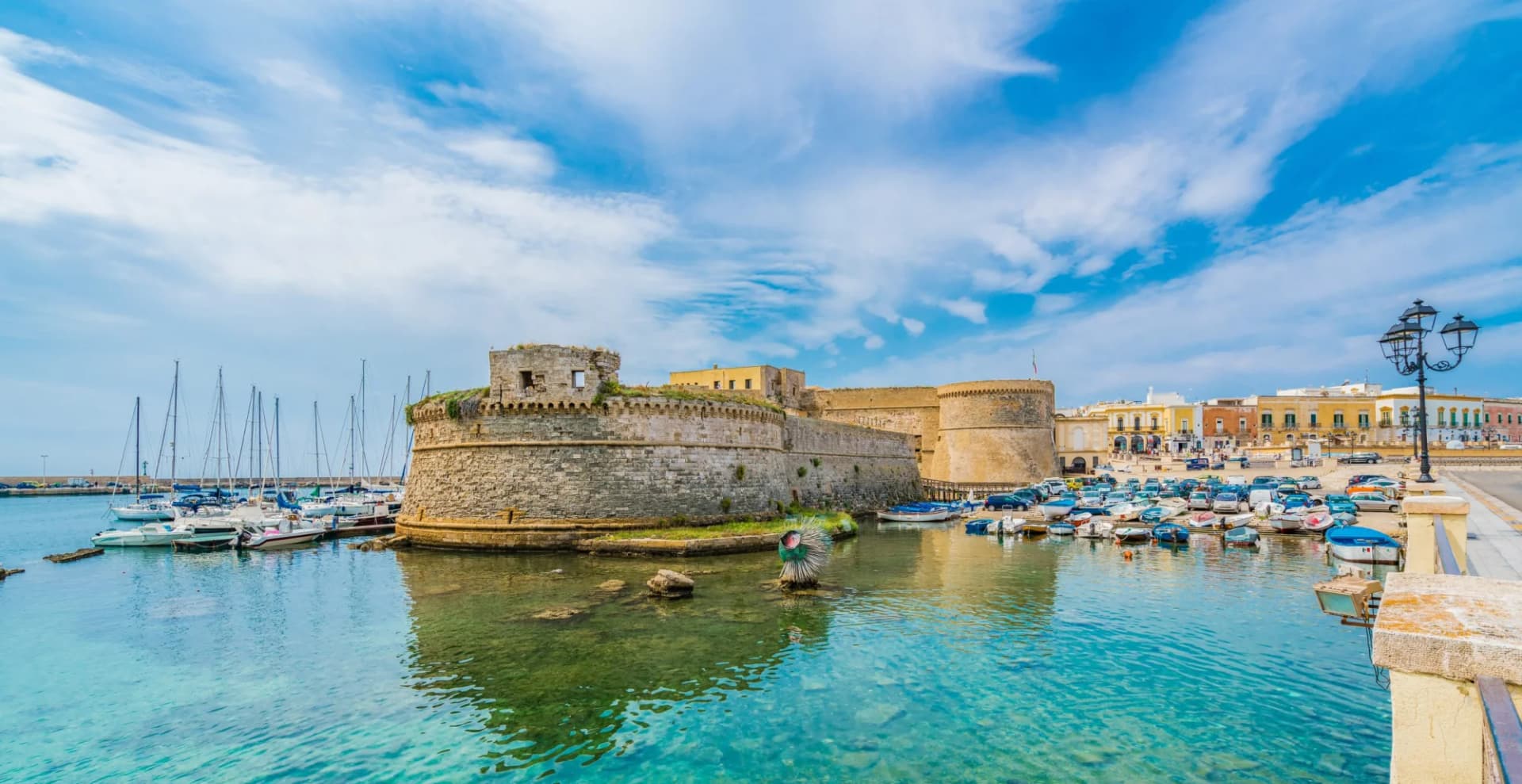 Stone fortress by harbor with clear turquoise water, docked boats, and colorful coastal town.